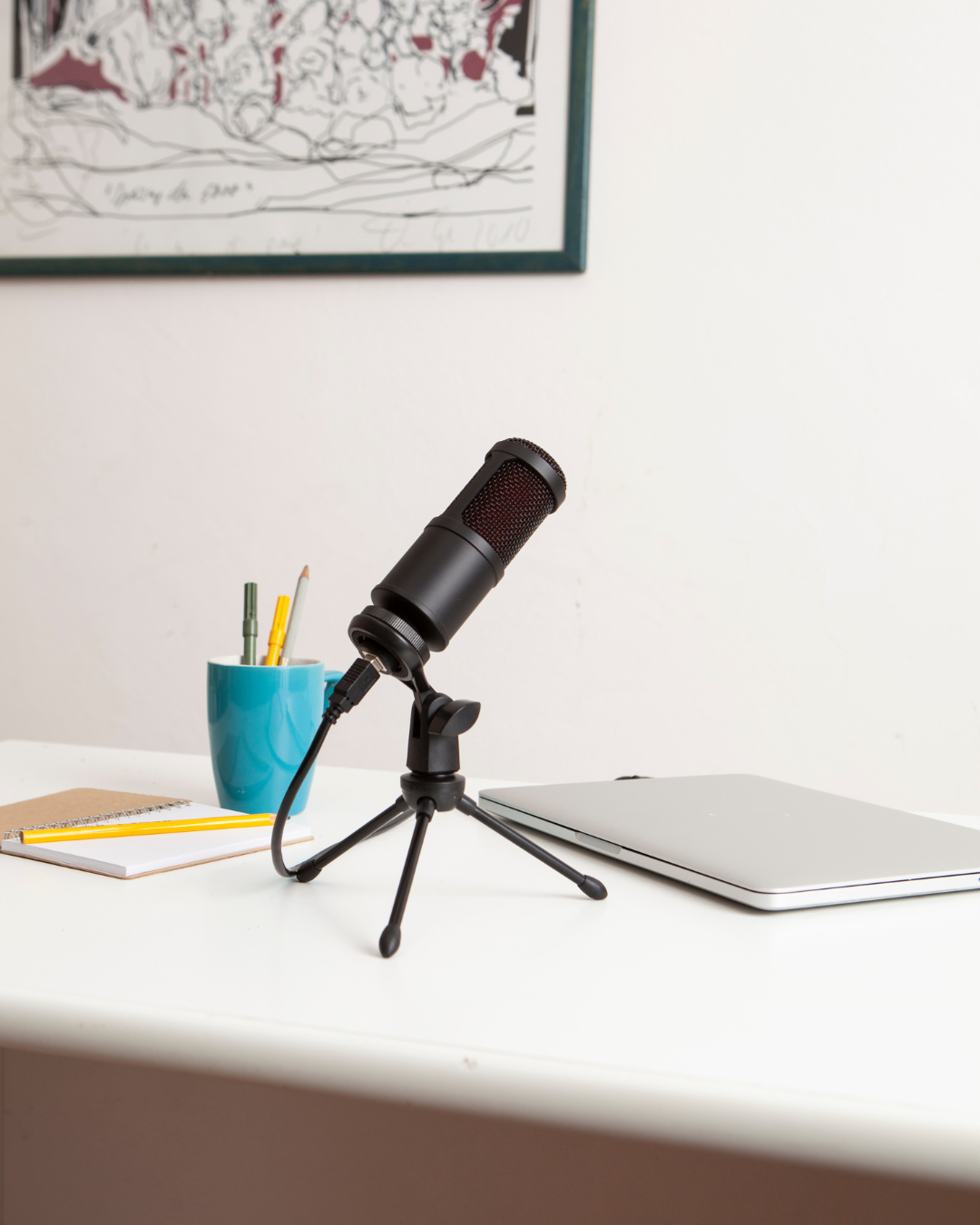 Image is of a desk with a black microphone, a laptop and a teal cup with pens and pencils in it