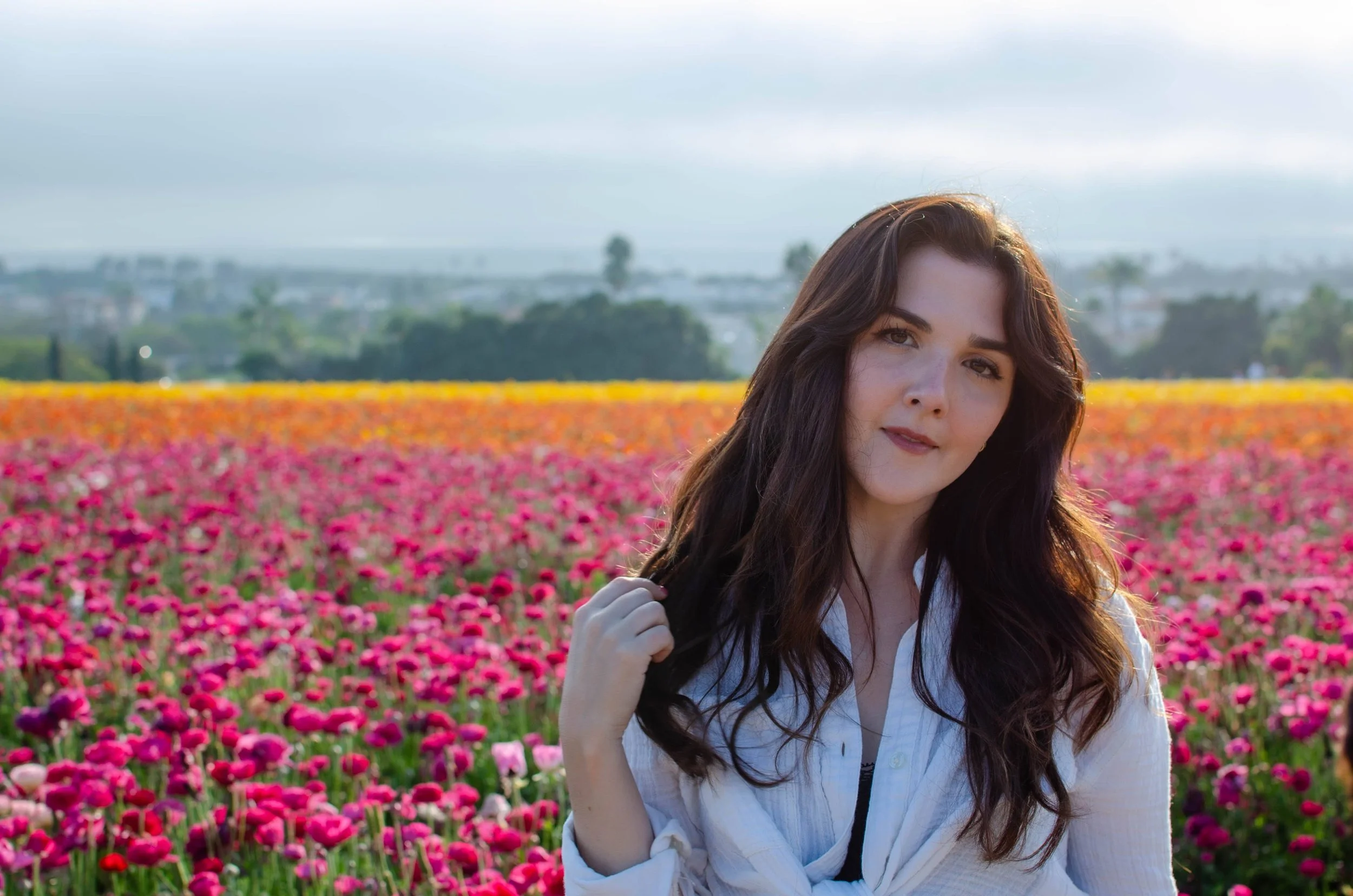 A woman with long dark hair standing in a colorful flower field with pink, red, and orange blooms, holding her hair, with trees and buildings in the background under a cloudy sky.