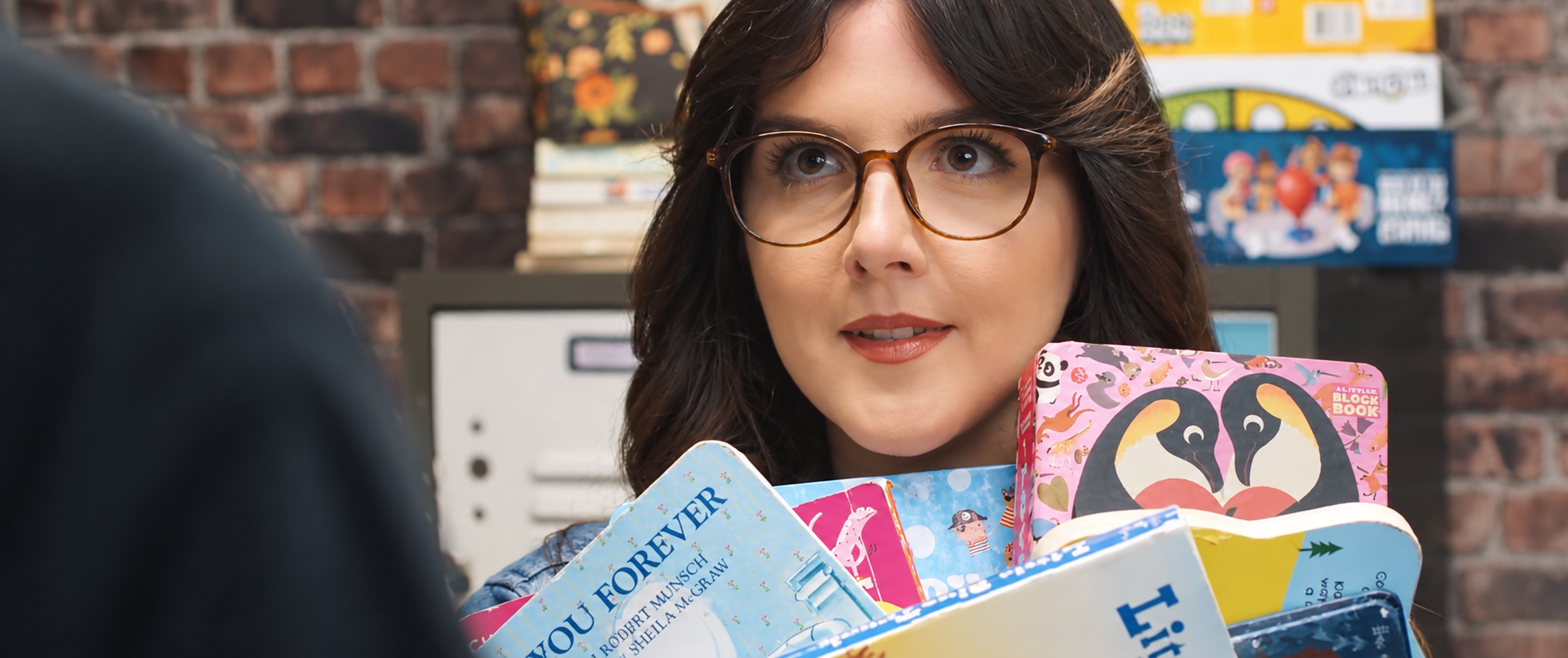 Woman with glasses holding a pile of children's books at a store counter, with a brick wall and shelves in the background.
