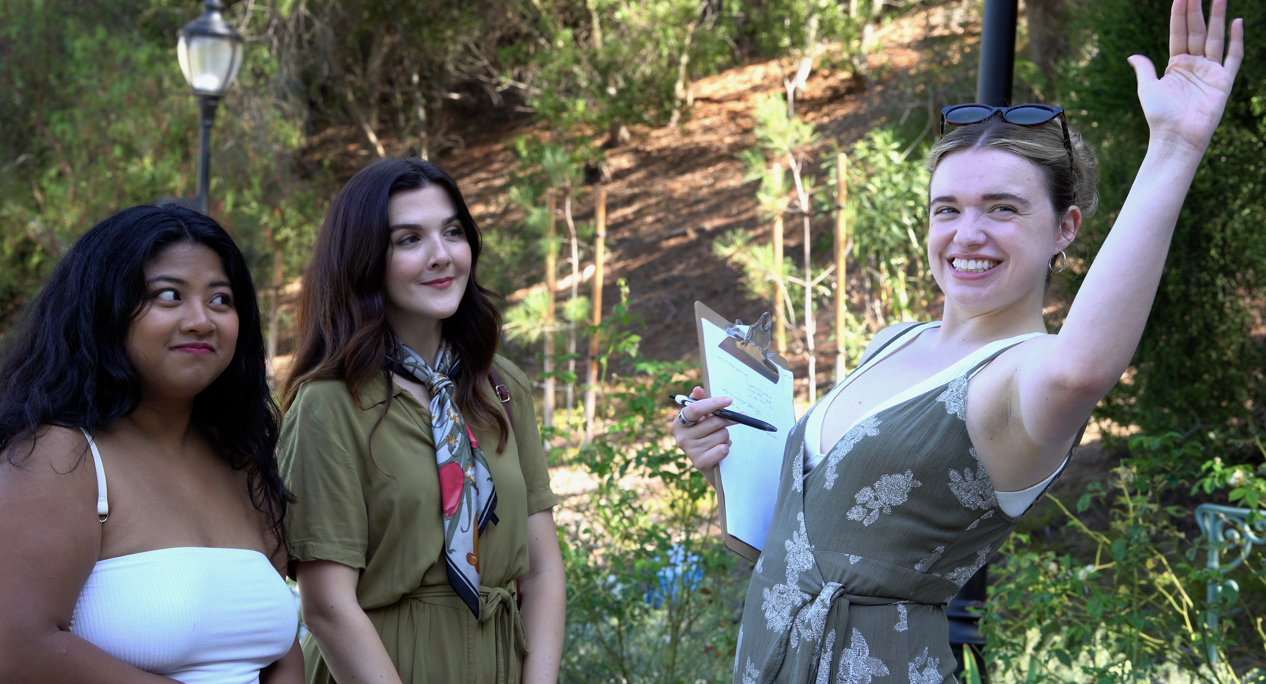 Three women in a garden, one smiling and waving, holding a clipboard, while the other two watch.