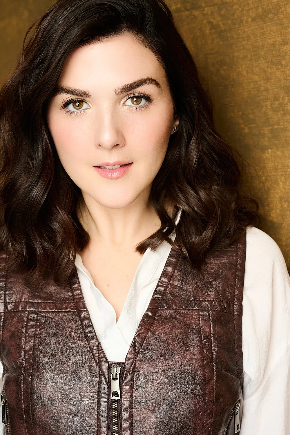 Close-up portrait of a woman with shoulder-length wavy brown hair, light skin, and hazel eyes, wearing a white shirt and a brown leather vest, standing in front of a wooden background.