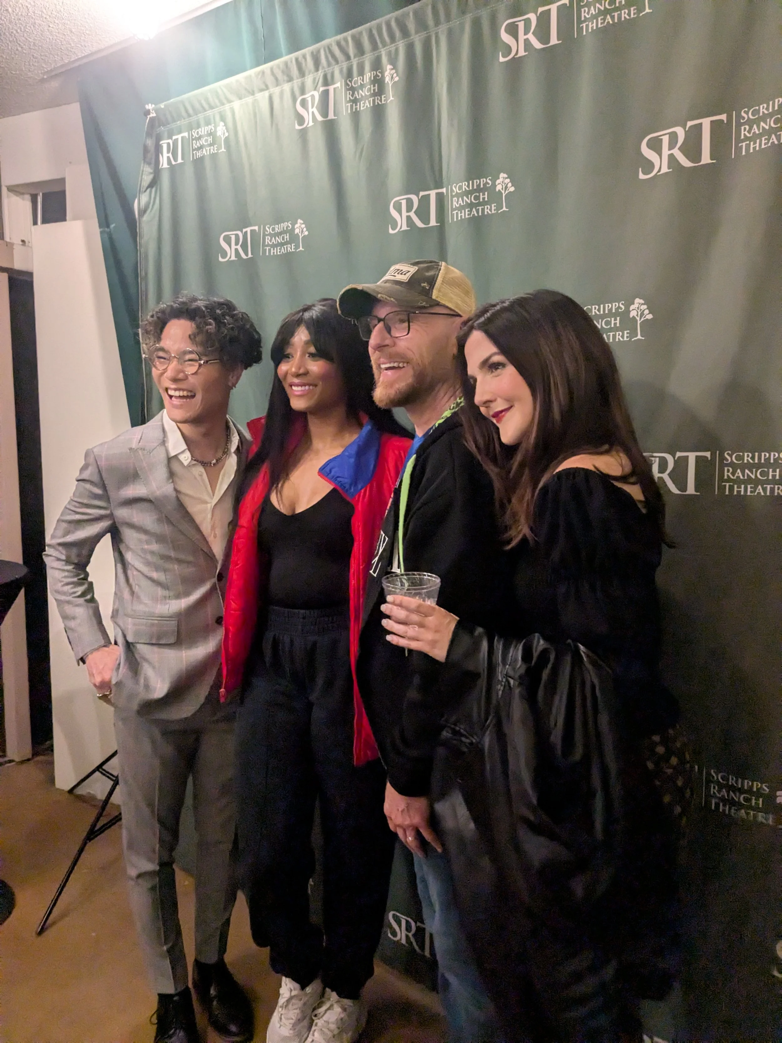 Four people standing together, smiling, in front of a green backdrop with 'SRT' and 'Scripps Ranch Theatre' logos. One person is holding a drink, and they are dressed casually and semi-formally, indicating a social event or gathering.