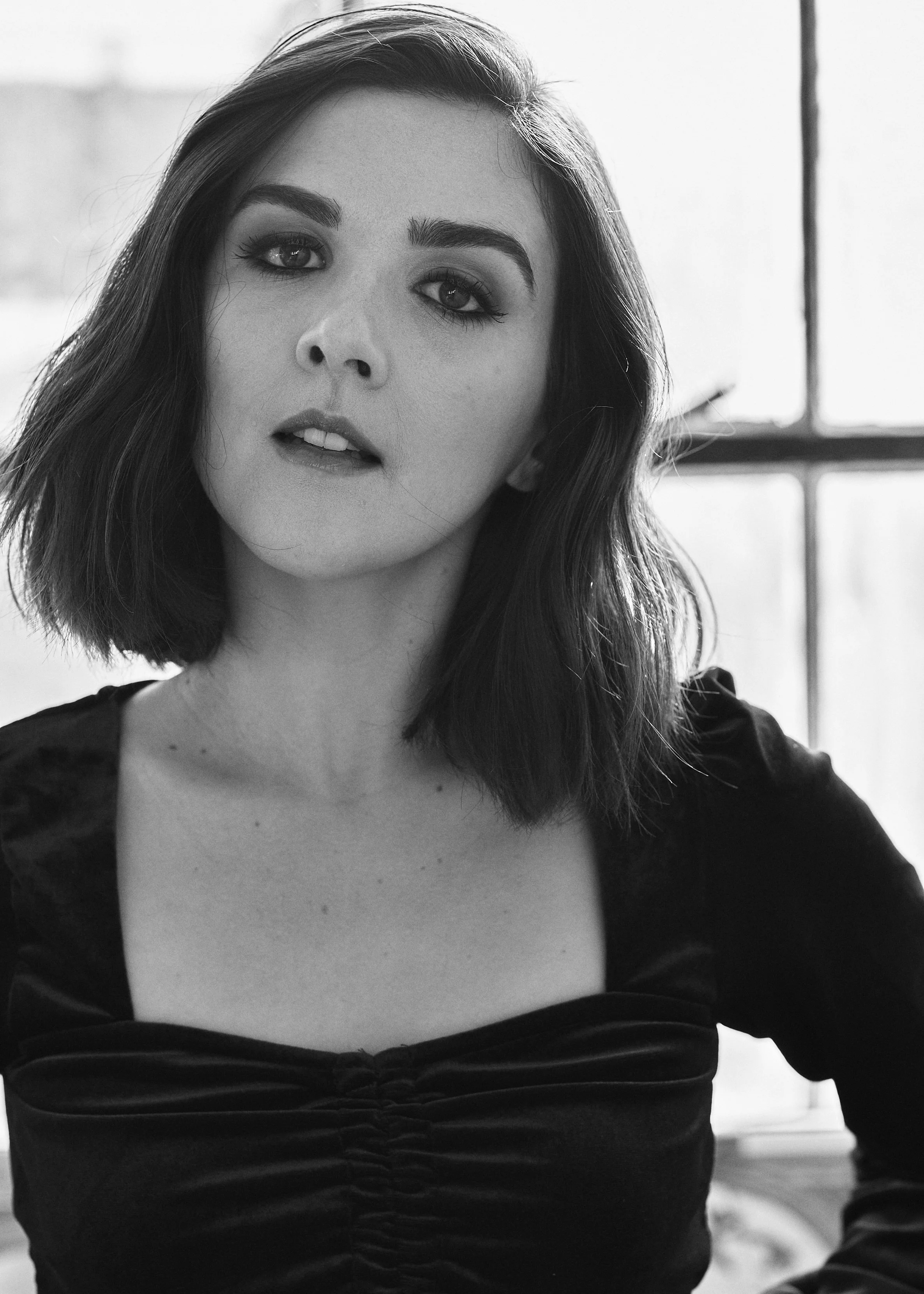 A black-and-white portrait of a woman with medium-length hair, wearing a black top with a sweetheart neckline, standing in front of a window with light coming through.