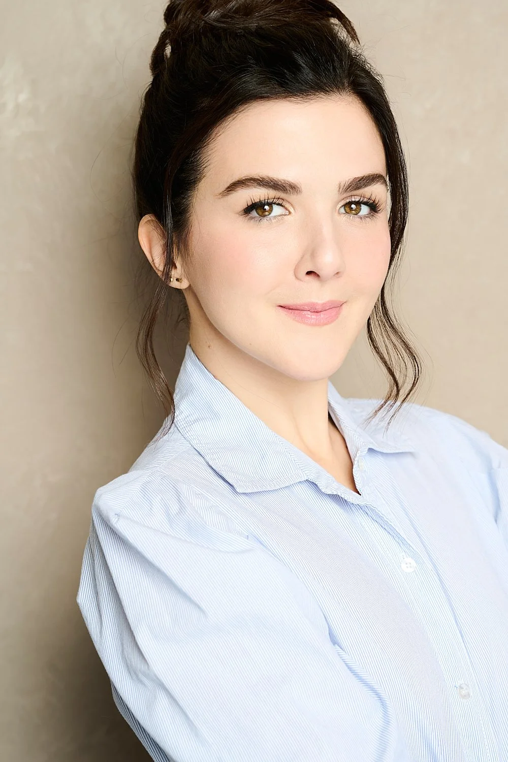 A young woman with dark hair styled in soft waves, wearing a light blue button-up shirt, standing against a plain, beige background.