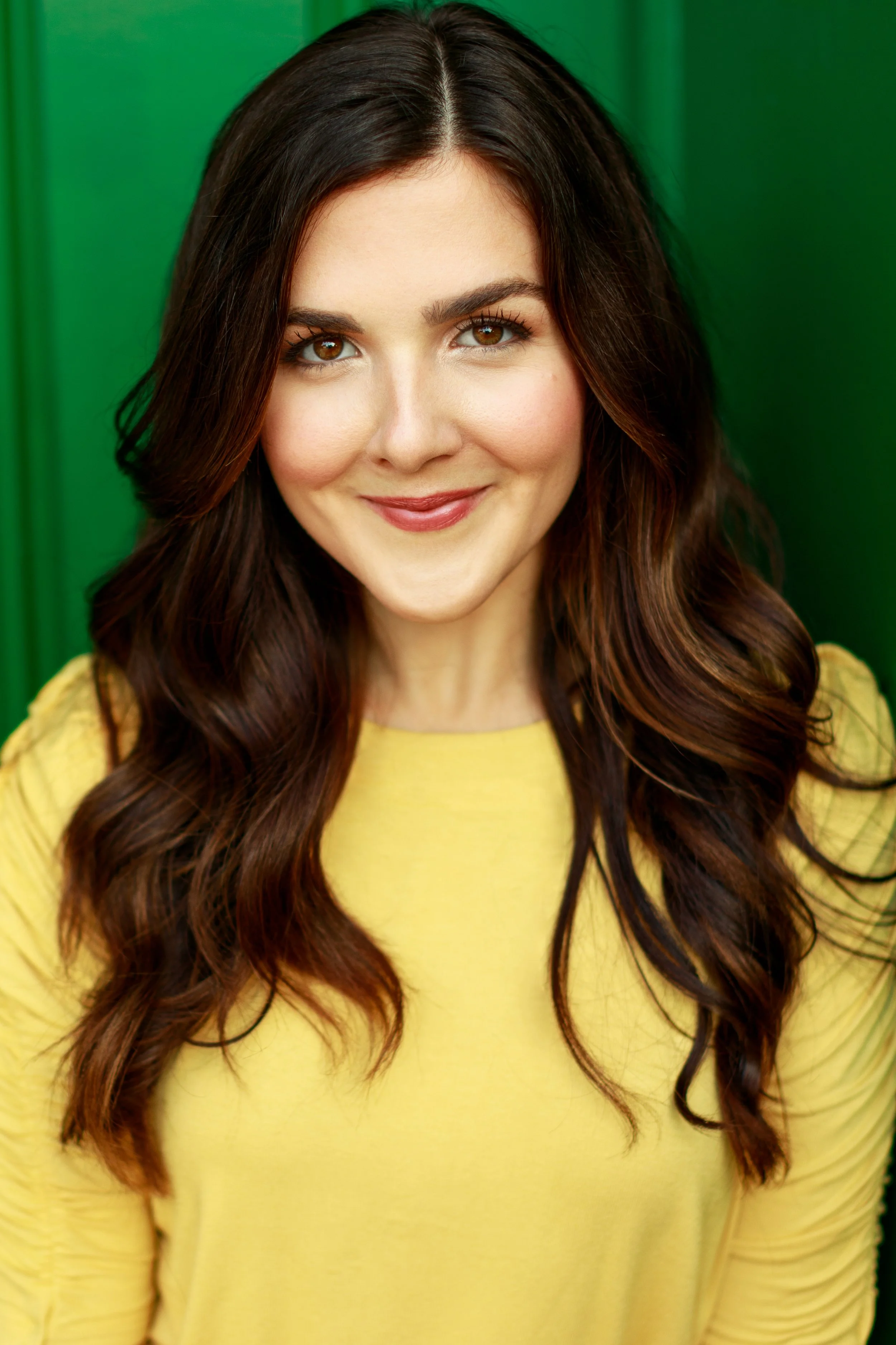 Close-up of a woman with long, wavy dark brown hair wearing a yellow top, smiling softly against a green background.