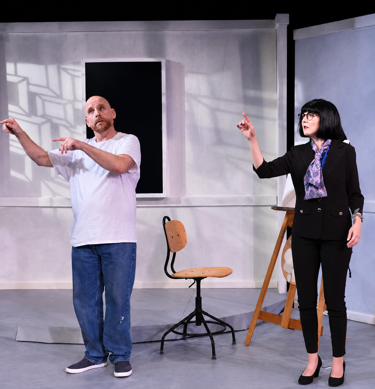 A man with a bald head and a beard wearing a white t-shirt and jeans is gesturing with his arms, while a woman with black hair, glasses, and a black suit with a patterned purple scarf is also pointing, both standing on a stage with a gray background 