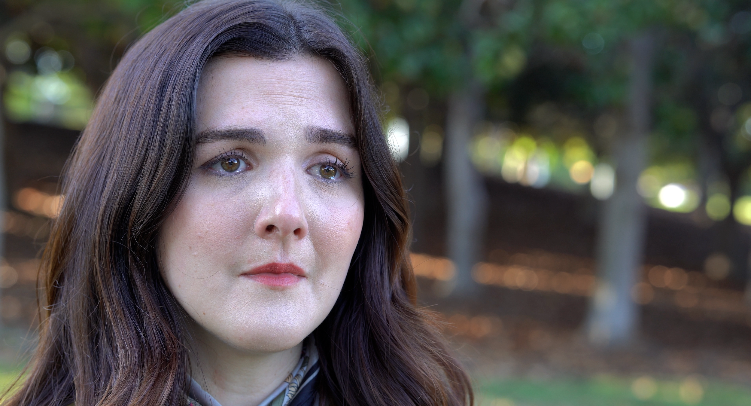 Close-up of a woman with long brown hair and hazel eyes outdoors in a park with trees and blurred greenery in the background.