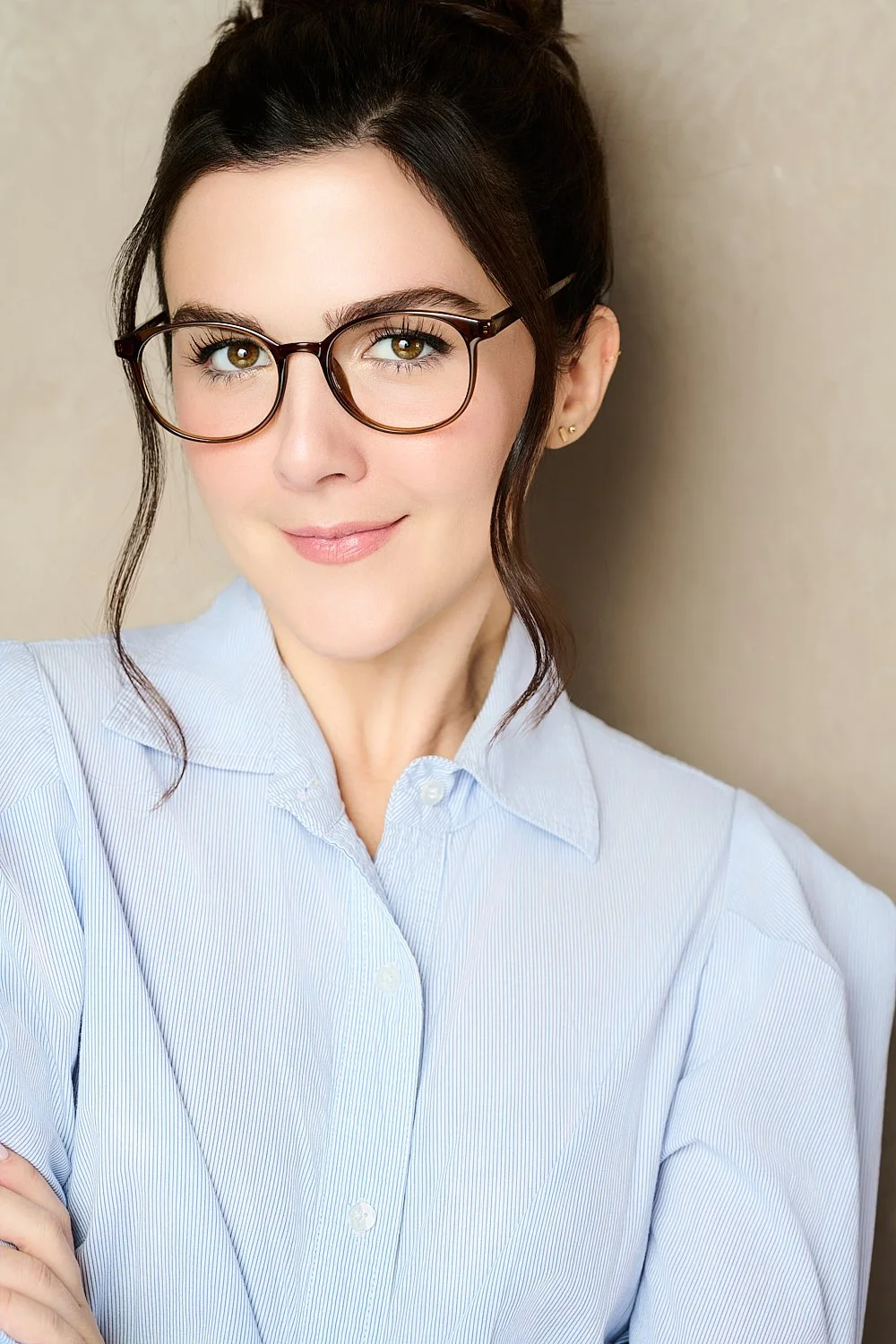 A woman with brown hair in a bun, wearing glasses and a light blue button-up shirt, smiling at the camera against a neutral background.