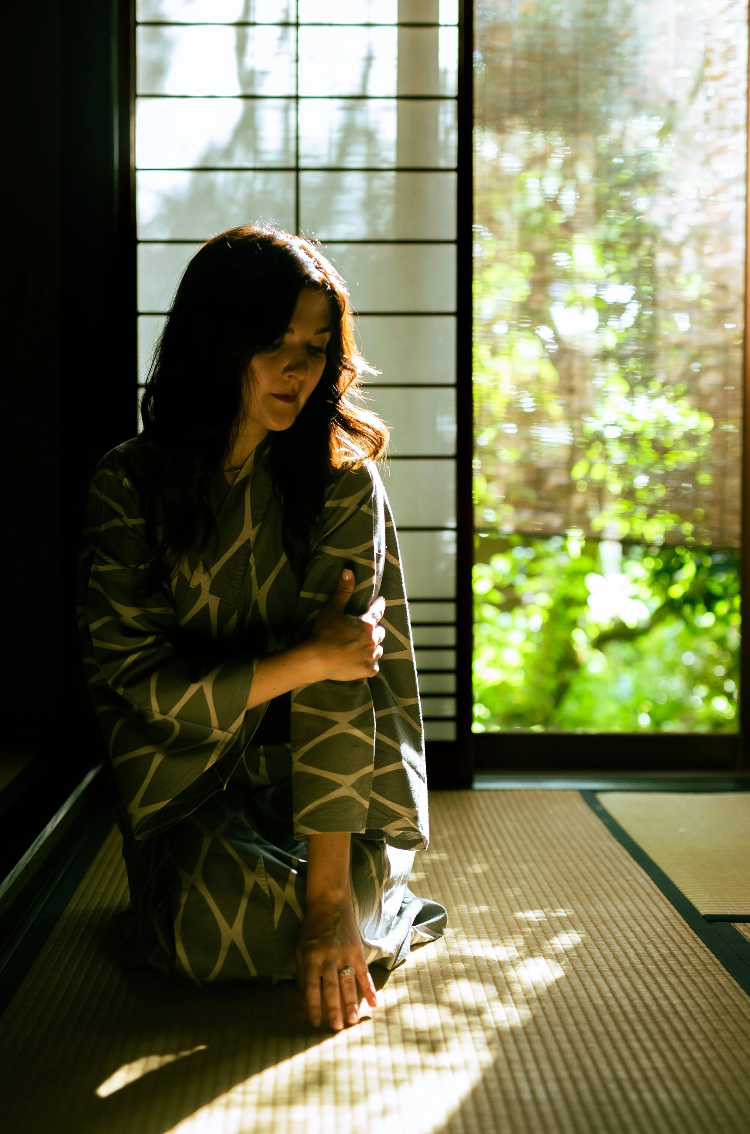A woman in traditional Japanese attire kneeling on tatami mat with sunlight streaming through shoji screens, casting shadows.