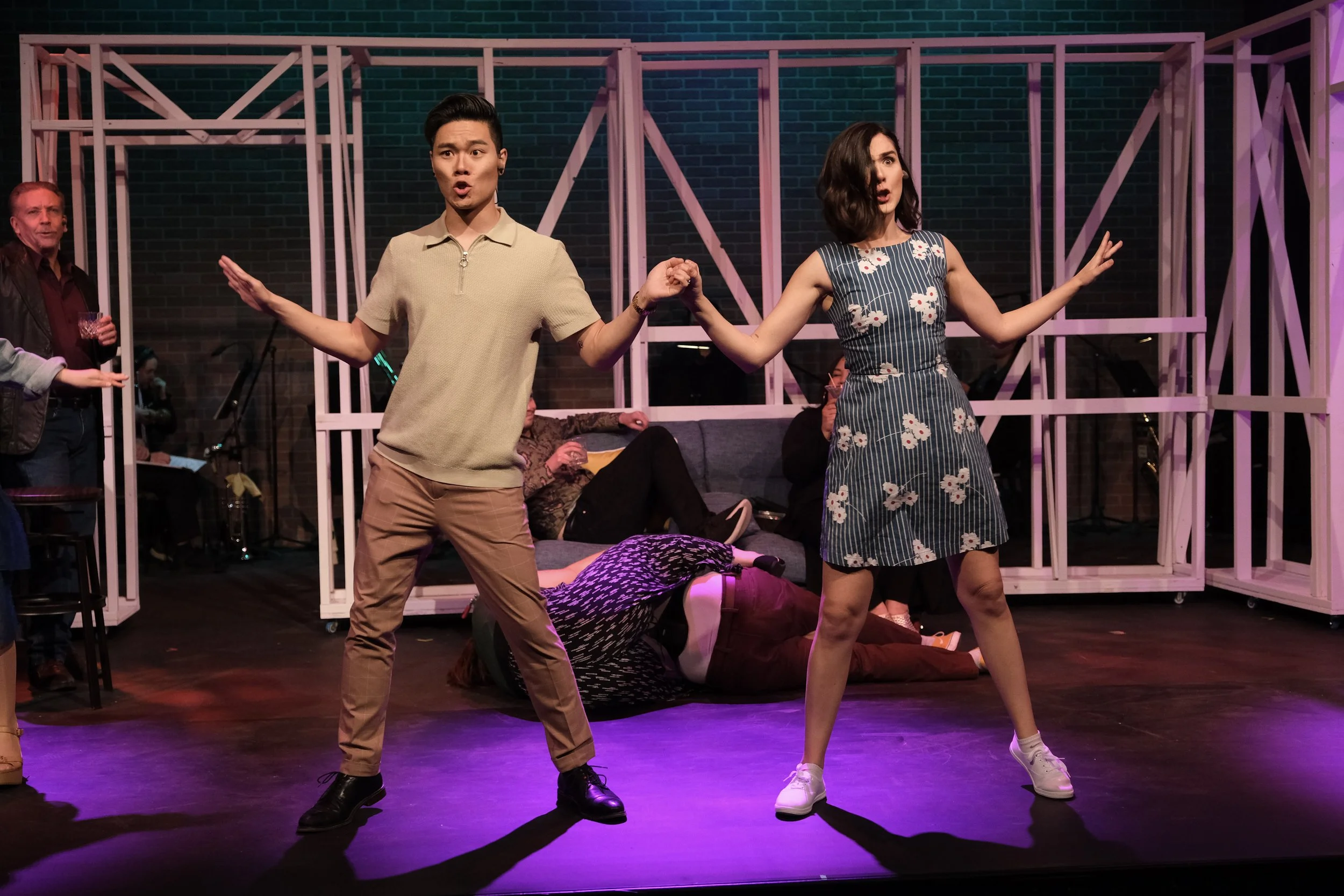 Two actors, a man and a woman, are performing on stage holding hands. The man has dark hair and is wearing a beige shirt and tan pants. The woman has dark wavy hair and is wearing a floral dress and white sneakers. There are other people and a person