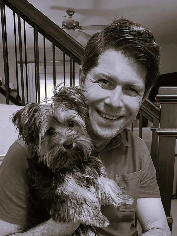 A man smiling while holding a small, furry dog indoors near a staircase.