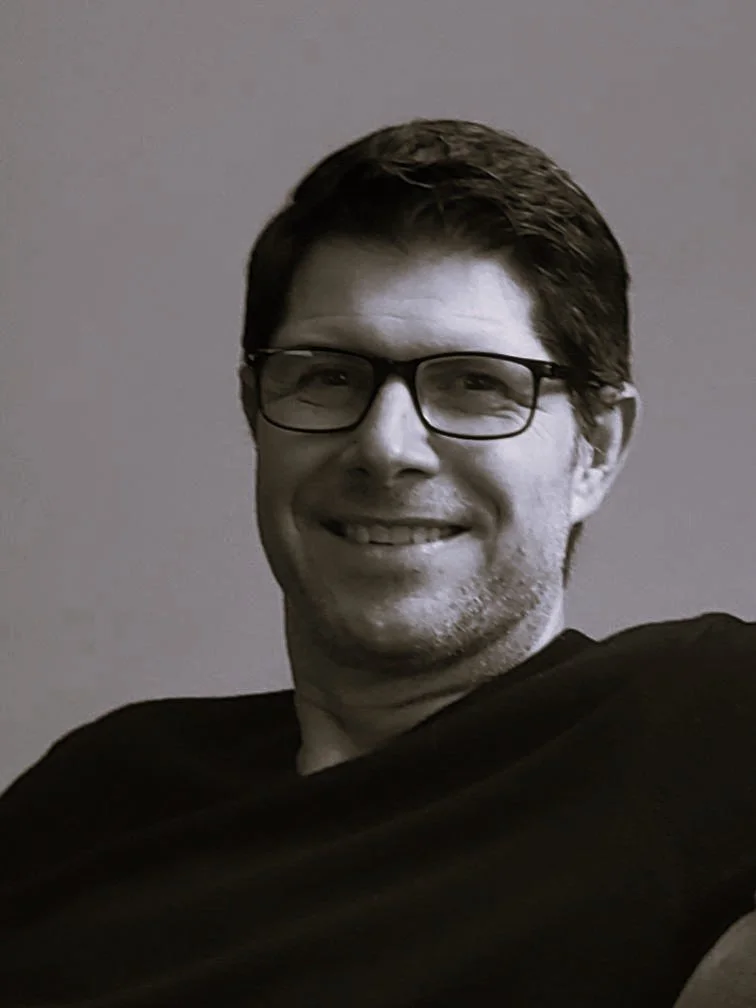 A smiling man, Heath Geiman, with glasses and dark hair, wearing a black shirt, sitting against a plain wall background.