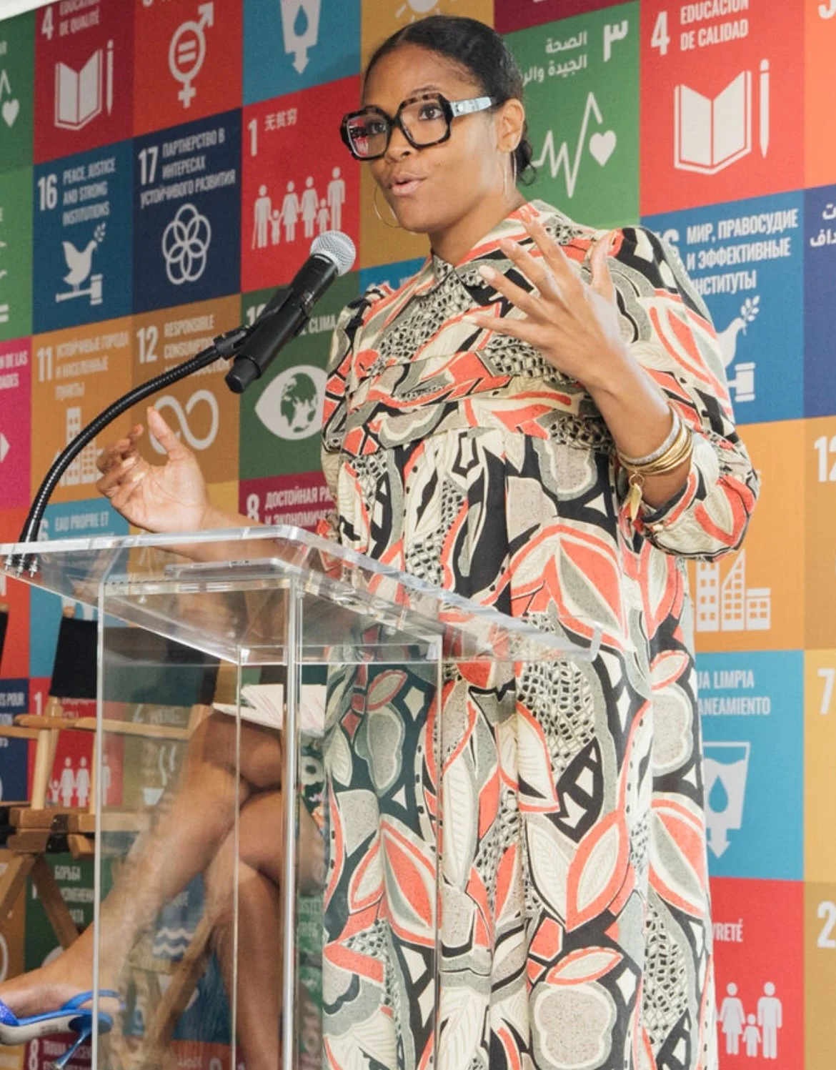 A woman speaking at a podium with a backdrop of colorful tiles featuring United Nations Sustainable Development Goals icons and text, wearing glasses and a patterned dress.