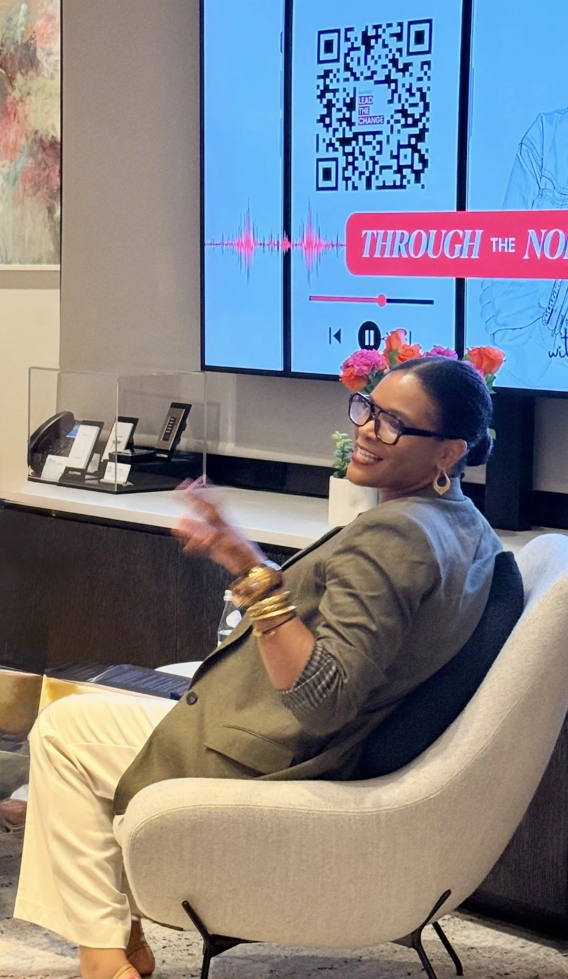 A woman with dark hair and glasses sitting in a modern beige chair, smiling and gesturing with her hand, in front of a large screen showing a QR code, waveform, and text that says 'Through the No' with a pink background and white text. There are flowers in a vase behind her and a display of phones on the counter.