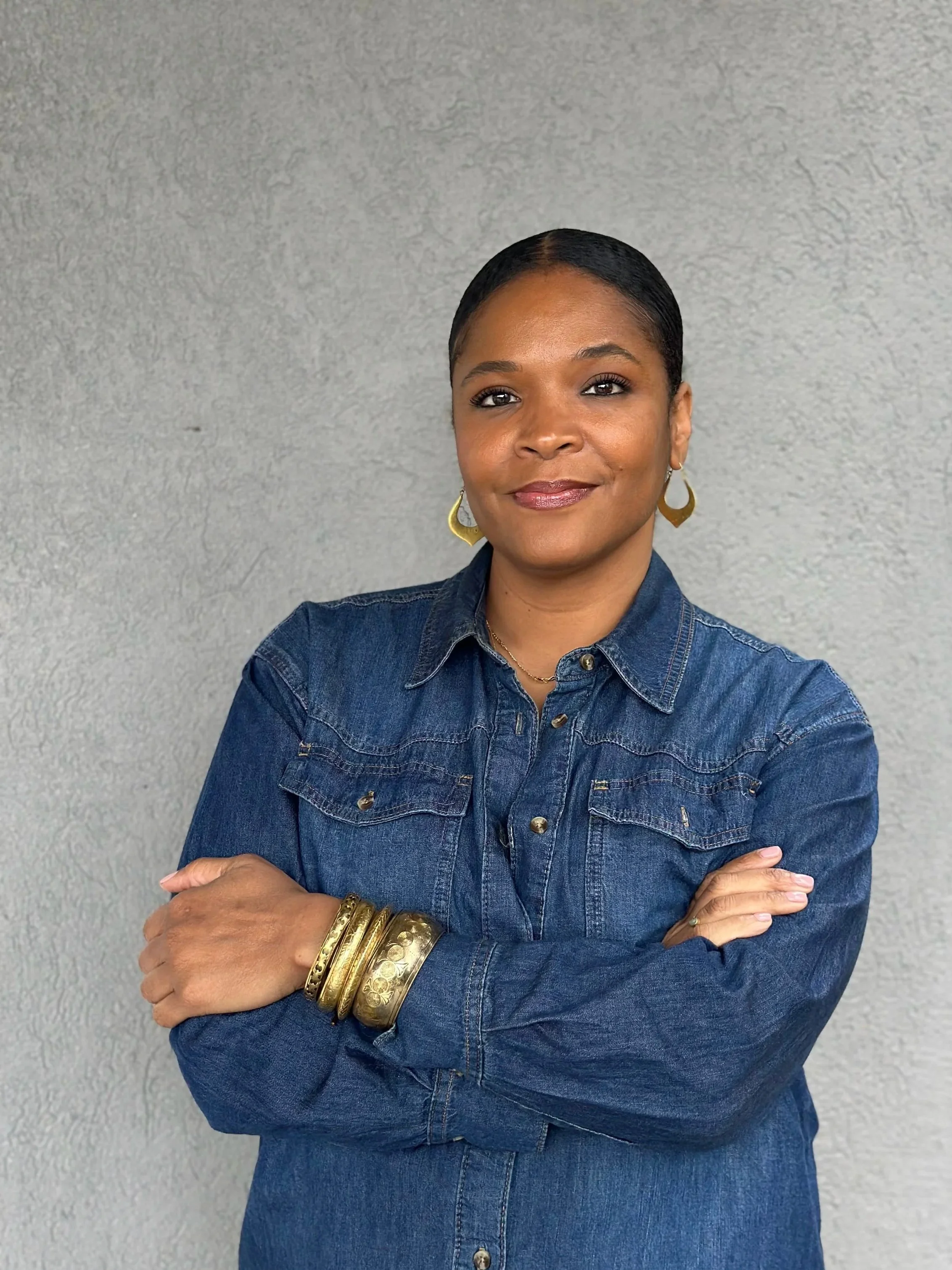 A woman with dark hair styled back, smiling with arms crossed in front of a plain gray wall. She is wearing a blue denim shirt, gold earrings, and multiple gold bangles.