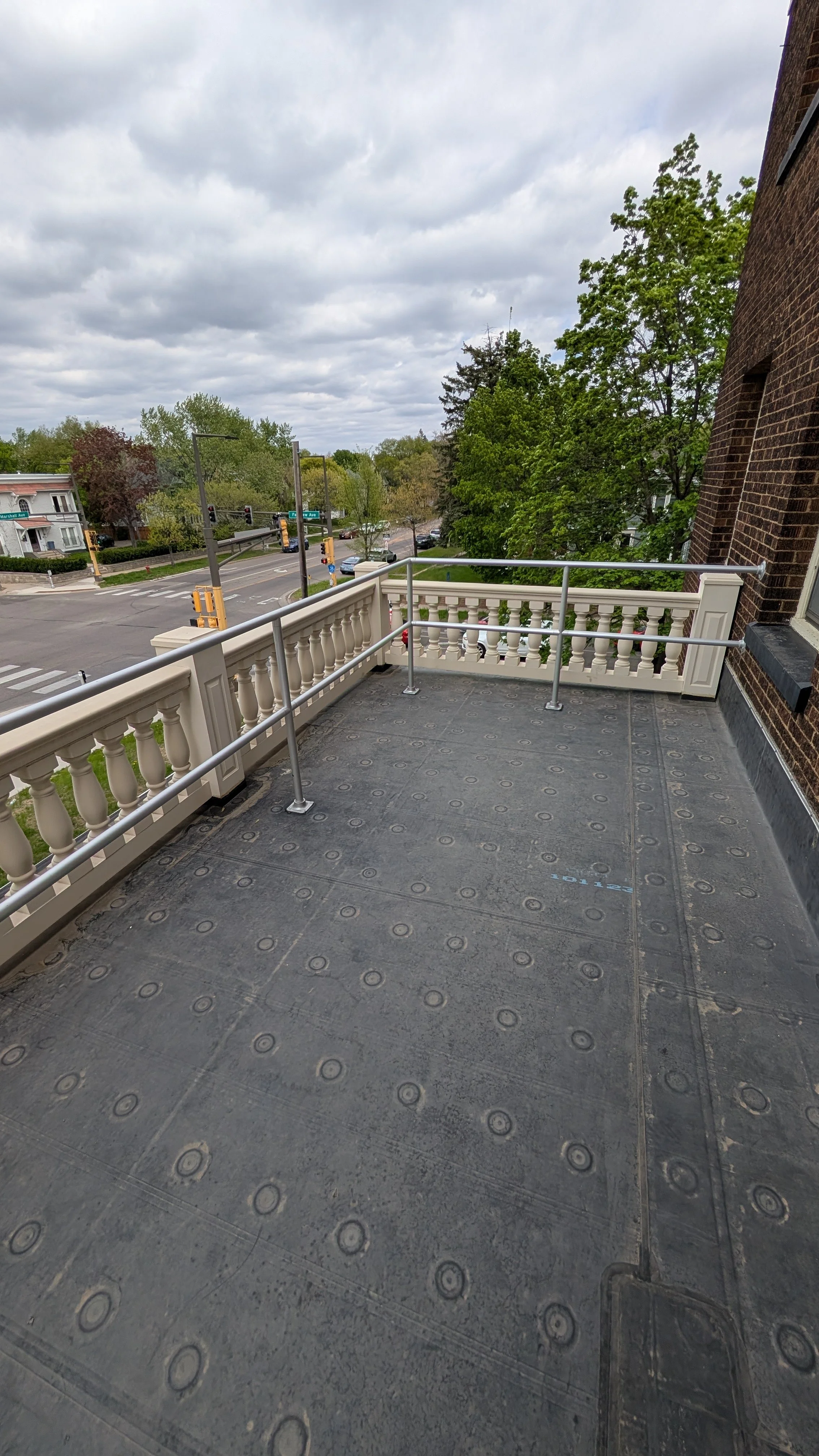 View from a balcony with a black surface, a white railing, and a city street with traffic lights, trees, and buildings in the background under a cloudy sky.