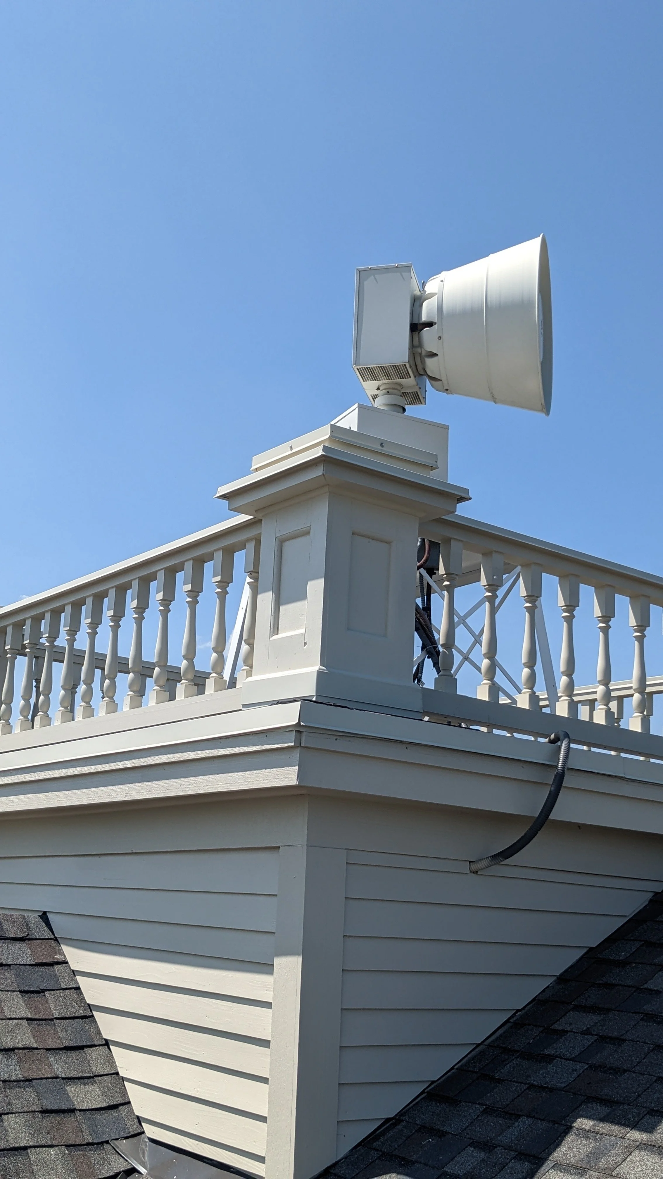 A white satellite dish mounted on the corner of a house rooftop with a white railing and beige siding, against a clear blue sky.
