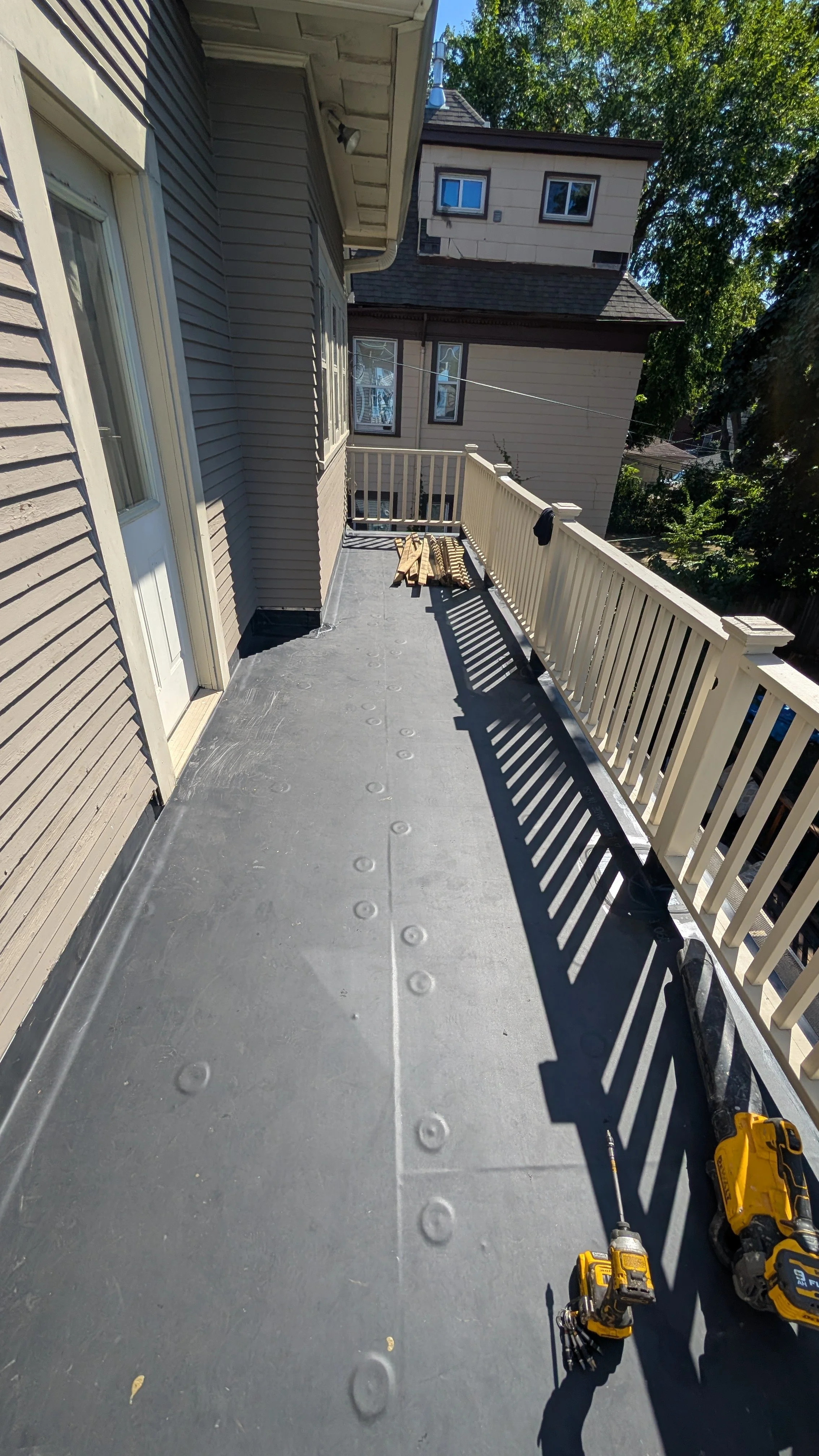 Empty balcony with a gray floor, beige railing, and beige siding on the house. There are two cordless drills on the floor, and a few wooden planks near the railing. The background shows trees and neighboring buildings.