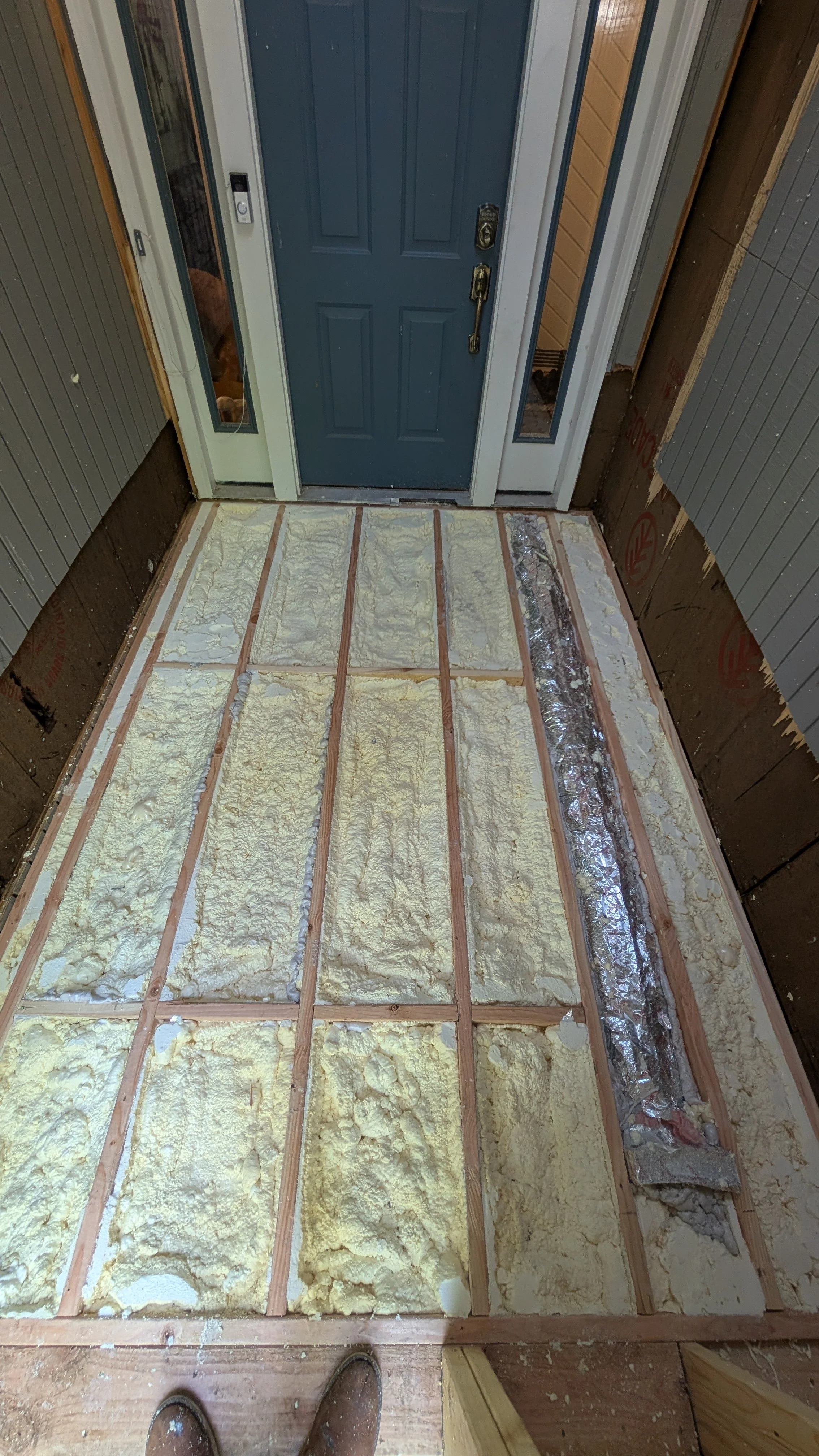 Entryway floor with exposed wooden subfloor and foam insulation, showing a blue door with side glass panels.