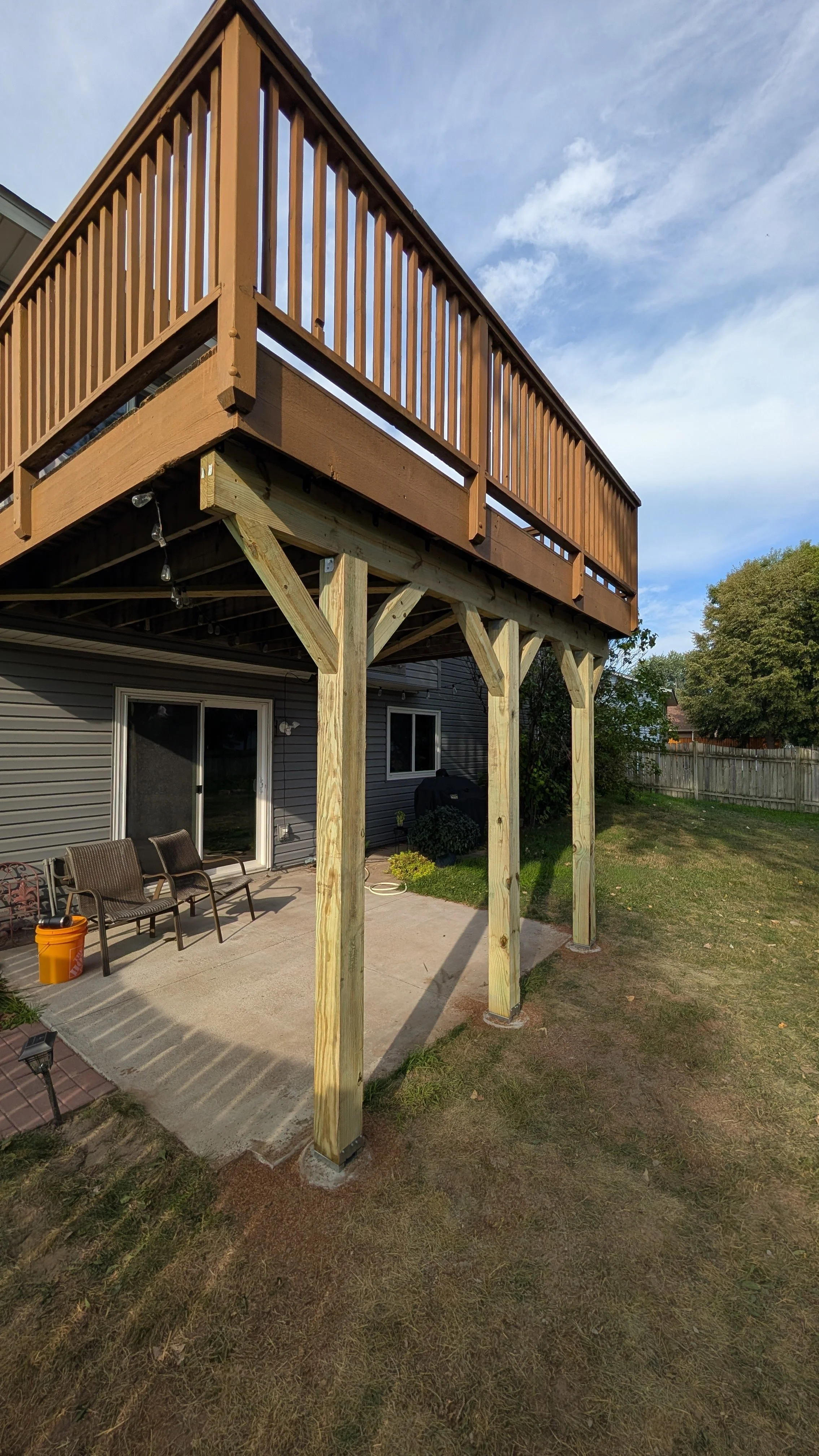 A backyard patio with a wooden deck extension being built, supported by vertical wooden posts, with outdoor chairs, a grill, plants, and a wooden fence in the background.