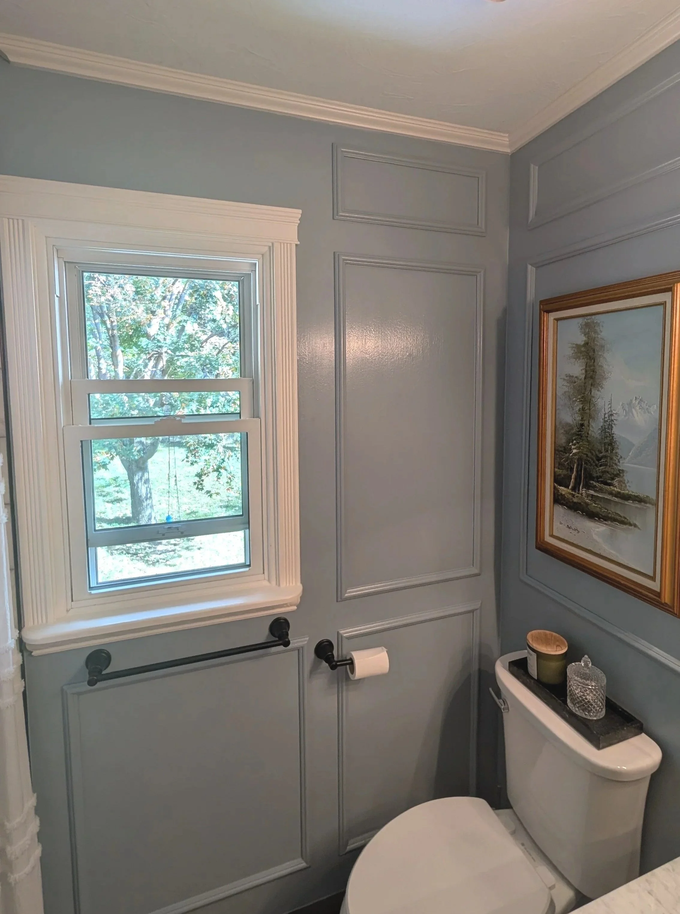 A bathroom featuring a light blue wall with decorative molding, a window with white trim, a bright outdoor scene visible through the window, a toilet with a black countertop, and decorative items including candles and a textured container on the countertop.