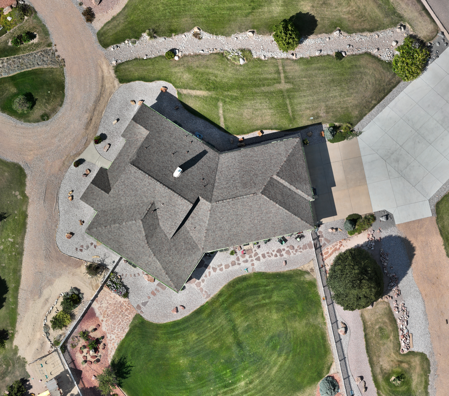 An aerial view of a backyard with a house, covered driveway, grassy yard, trees, pathways, and garden areas.