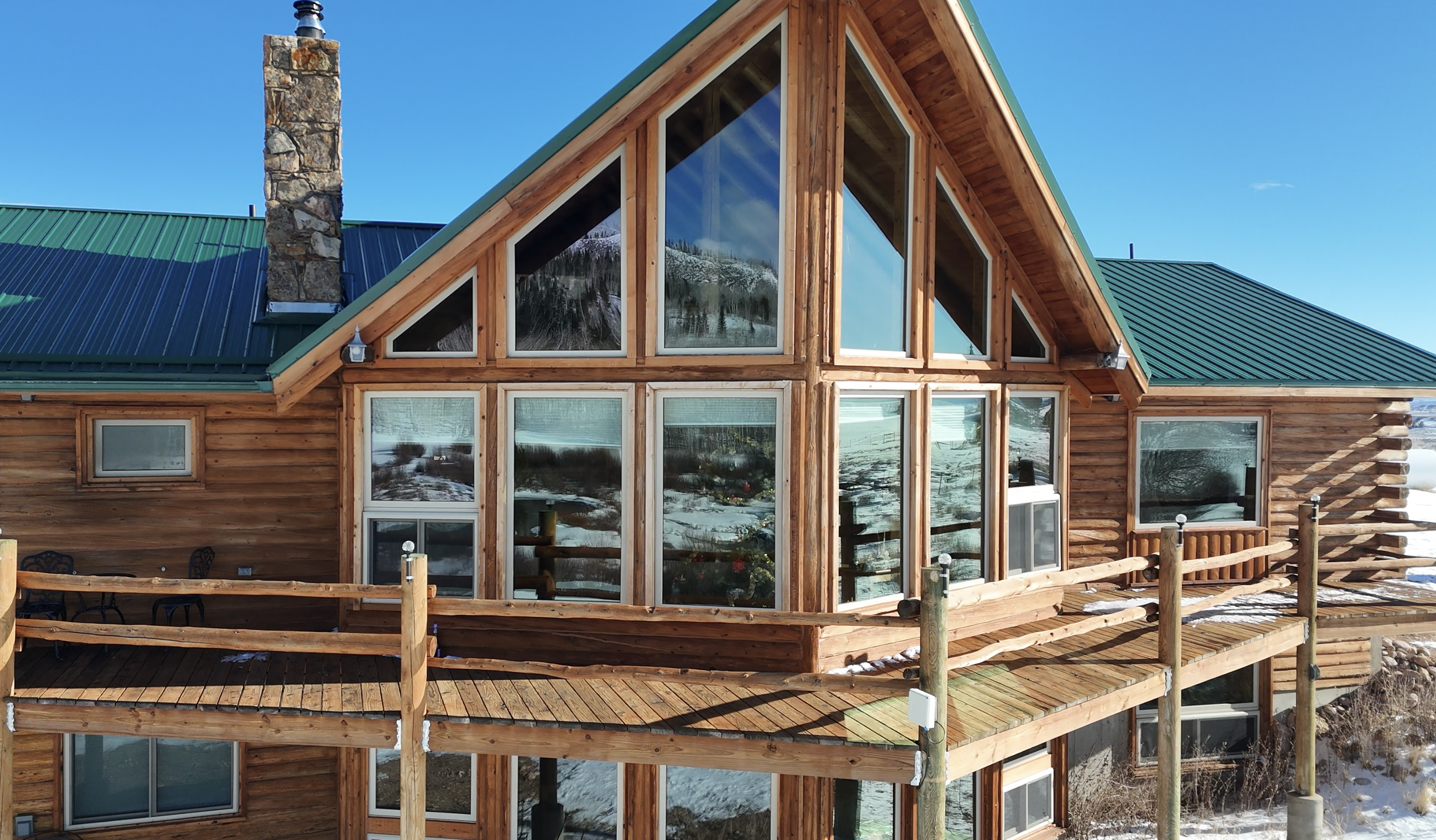 Wooden house with large glass windows and a green metal roof, with snow on the ground and mountains reflecting in the windows.
