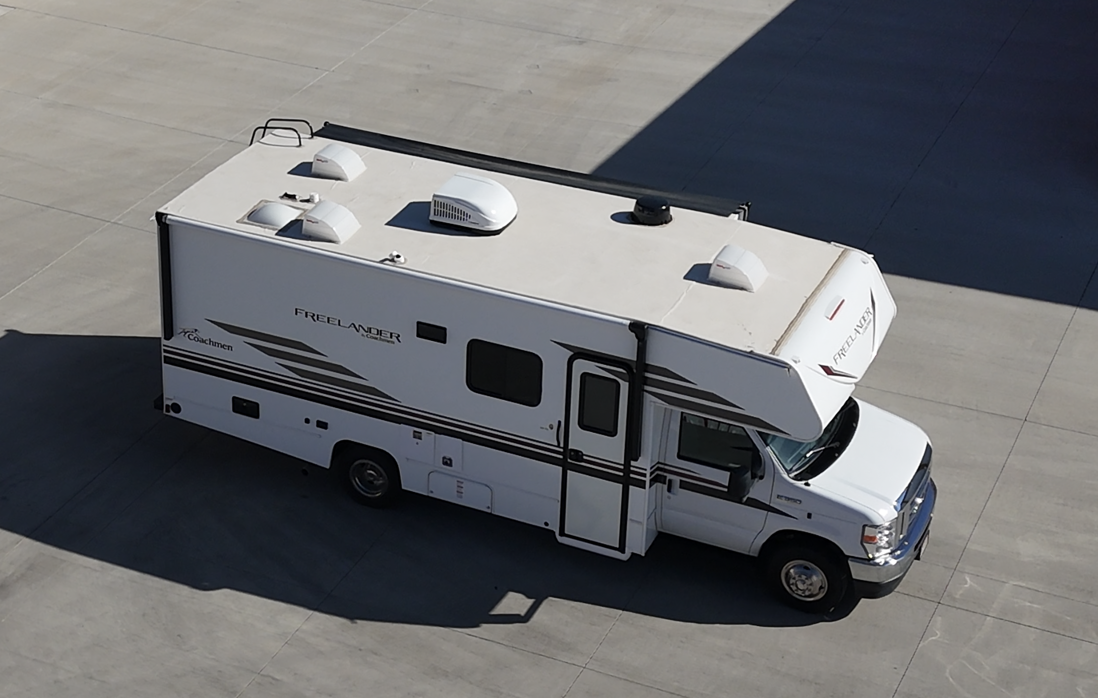 A white recreational vehicle (RV) with black and gray graphics, parked on a large concrete surface. The RV has multiple roof vents, a satellite dish, and an awning. One side of the RV features the brand name 'Freelander' and the model name 'Coachmen'