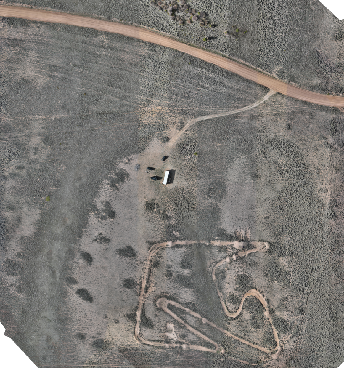 An aerial view of a rural landscape showing a winding dirt trail, sparse vegetation, a building, and several vehicles near the building.