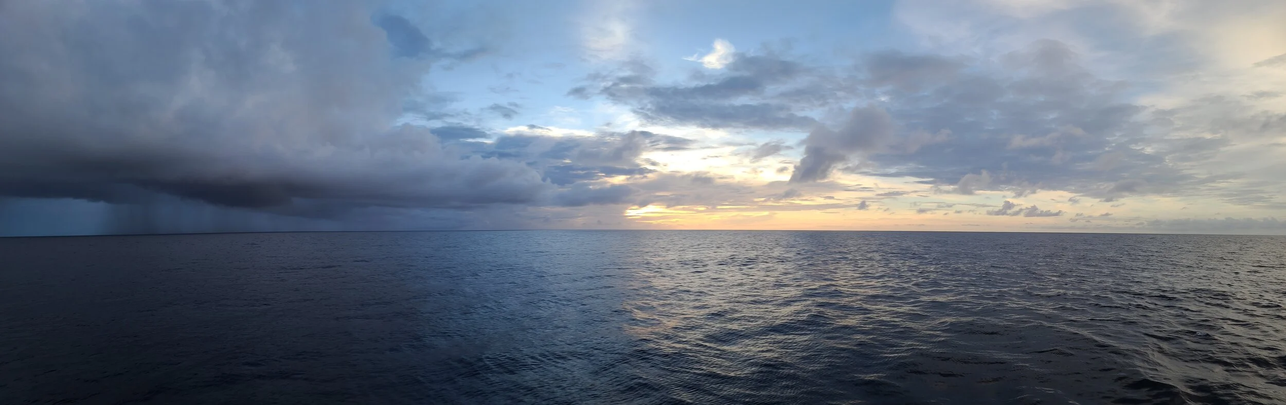 Ocean with a cloudy sky at sunset, including dark rain clouds on the left and lighter clouds with a few blue patches on the right.