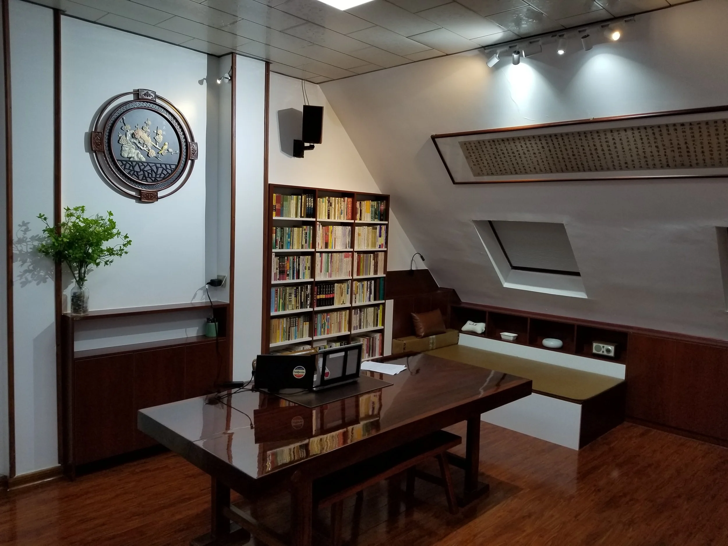 A cozy attic home office with a sloped ceiling, wooden accents, a bookshelf filled with books, a desk with a laptop and papers, and a built-in bench with cushions. There is a round wall clock, a small plant, and skylight windows on the ceiling.