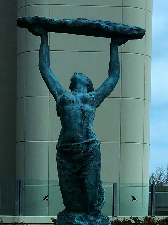 A bronze statue of a woman holding a large flat object above her head, standing outdoors against a modern building.