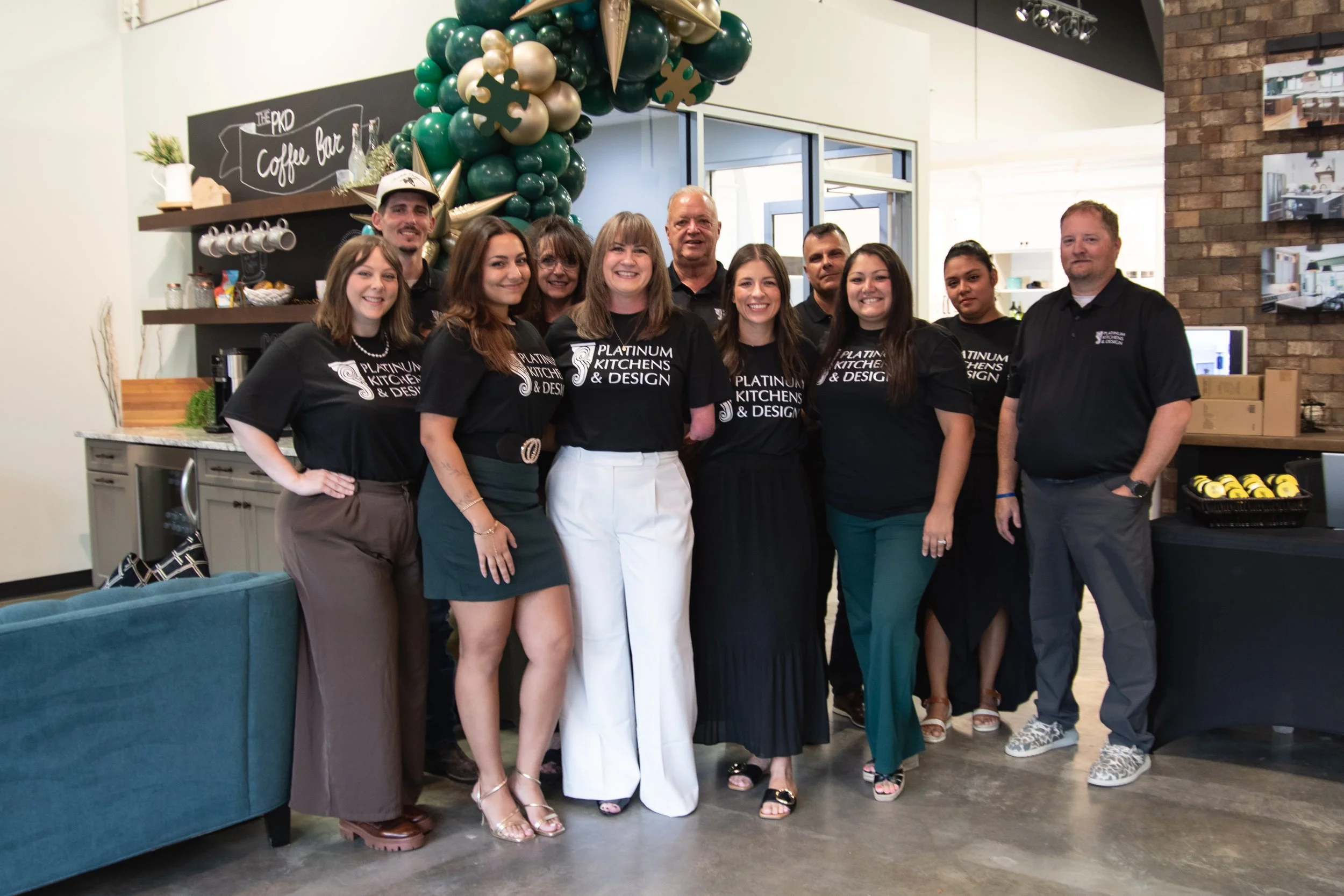 Platinum Kitchens & Design team posing beneath a green and gold balloon arch in a showroom.