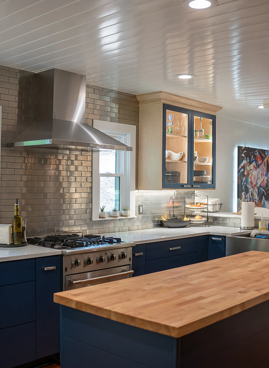 Modern kitchen with navy cabinets, wood island, stainless steel range hood, gas stove, subway tile backsplash, and glass-front shelving
