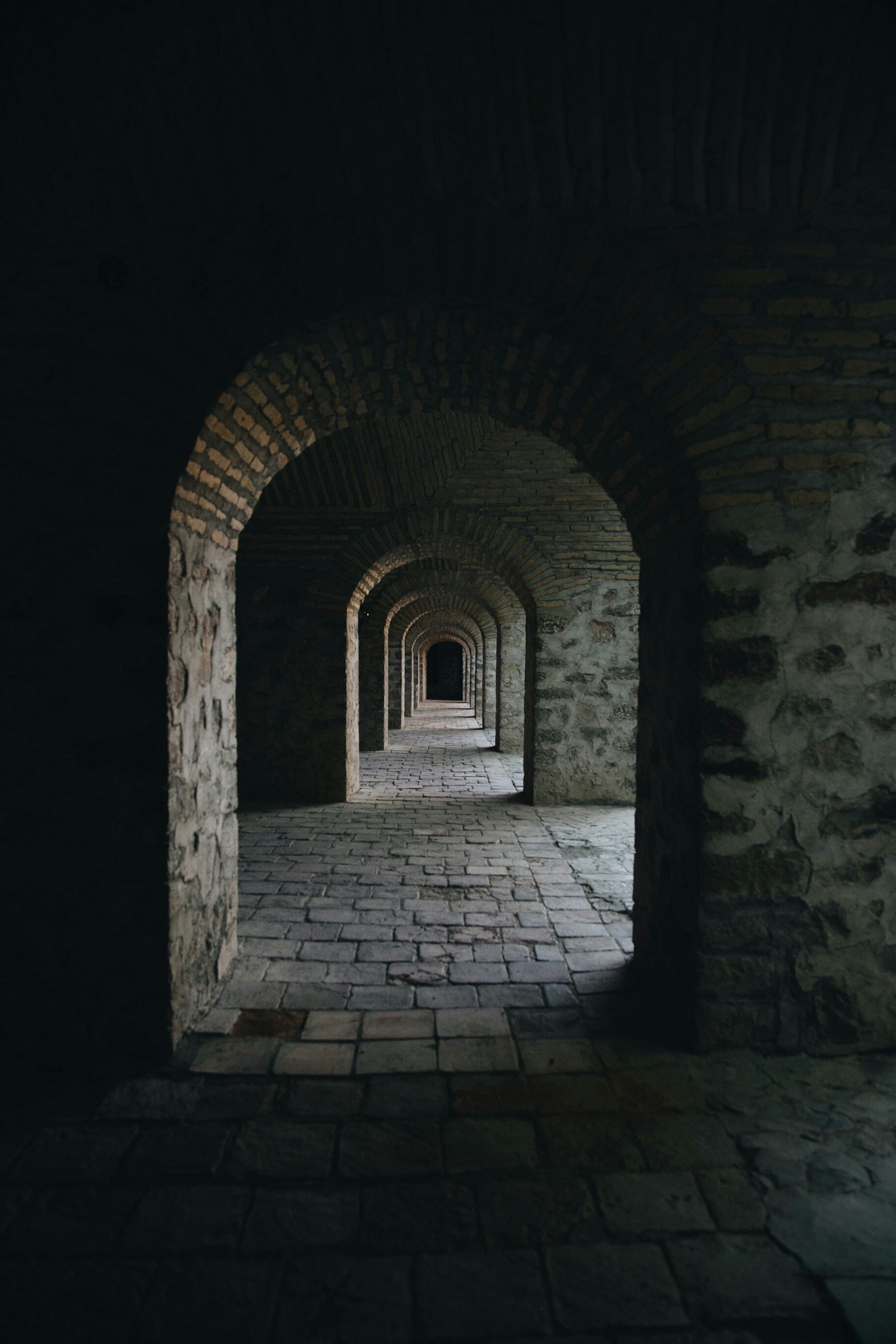 A series of stone archways in a dimly lit corridor, with cobblestone flooring and textured stone walls, creating a tunnel-like effect.