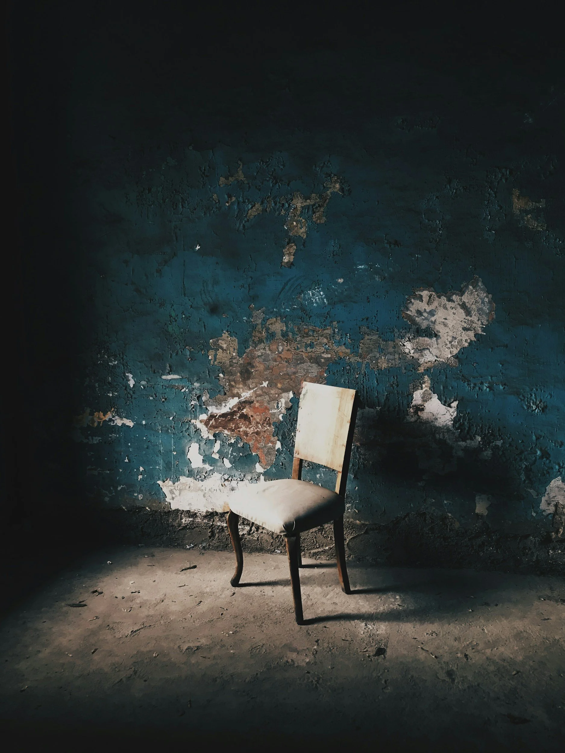 An old plain chair with a worn cushion, situated in a dark, dilapidated room with peeling blue paint on the wall.
