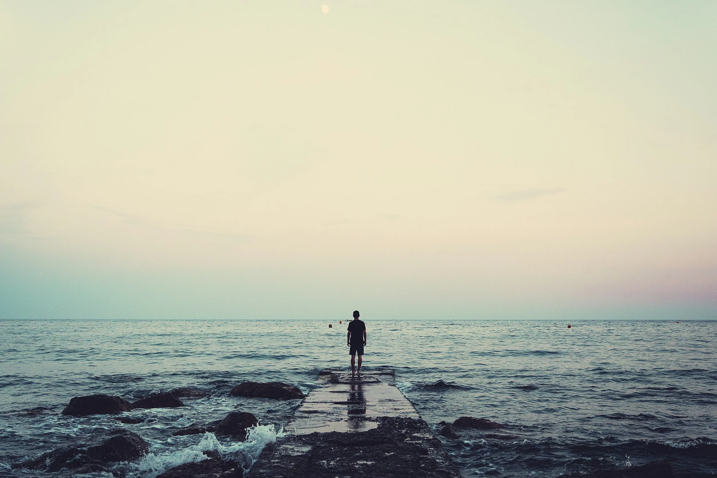 Person standing on a rocky pier looking out over the ocean at sunset.