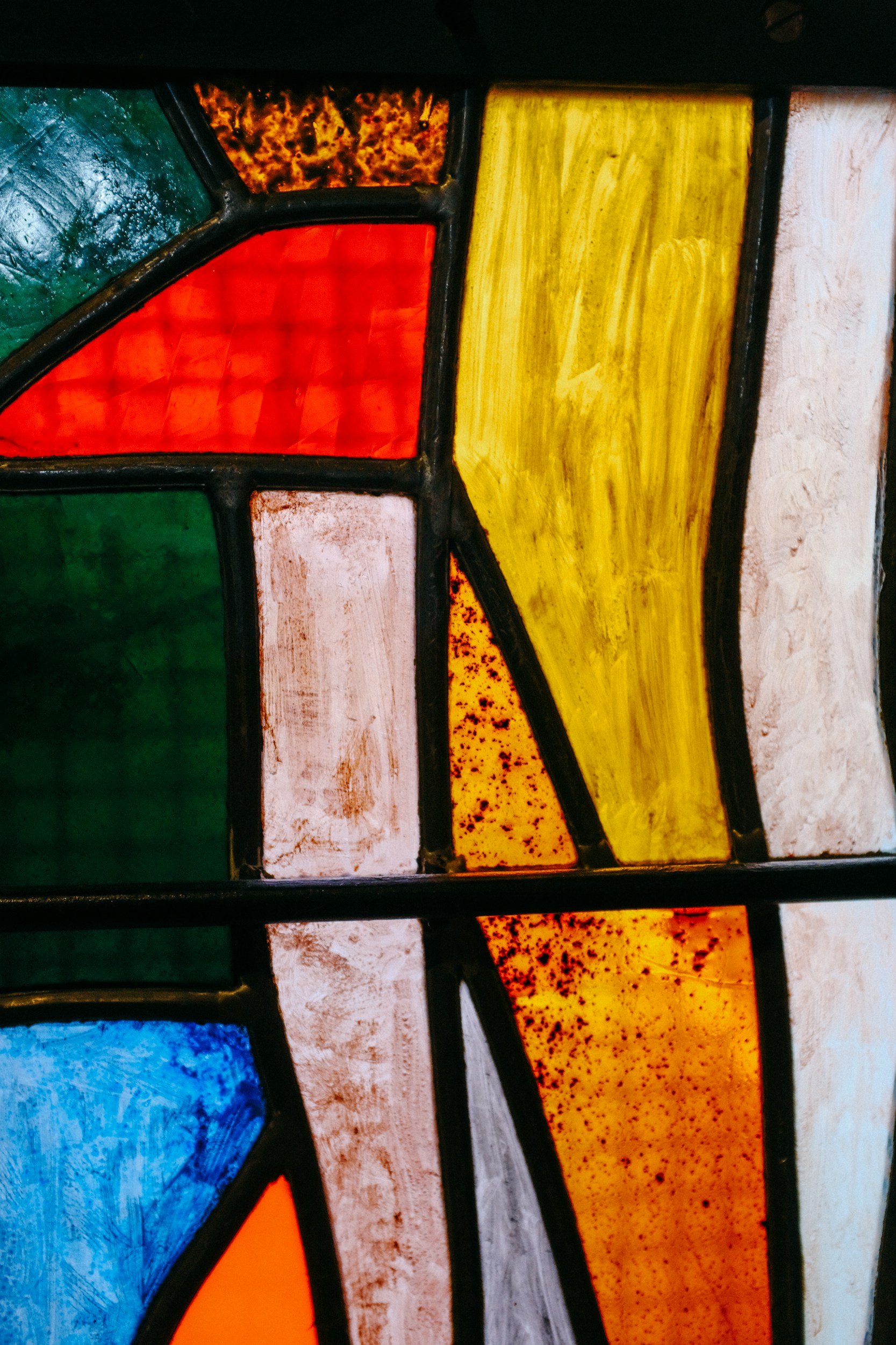 Close-up of a stained glass window with colorful geometric shapes, including green, red, yellow, orange, white, blue, and textured black glass, separated by black leading lines.