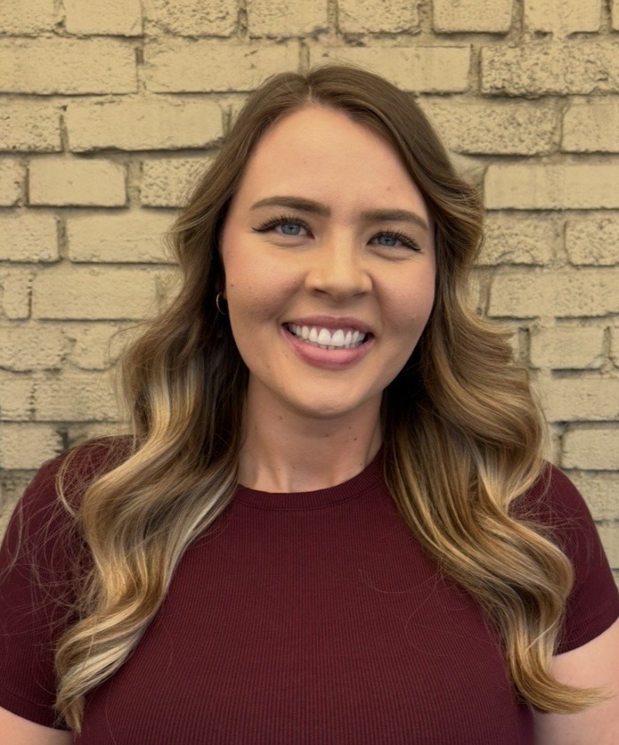 A young woman with long, wavy brown hair and blue eyes, smiling while standing in front of a beige brick wall, wearing a dark red t-shirt.