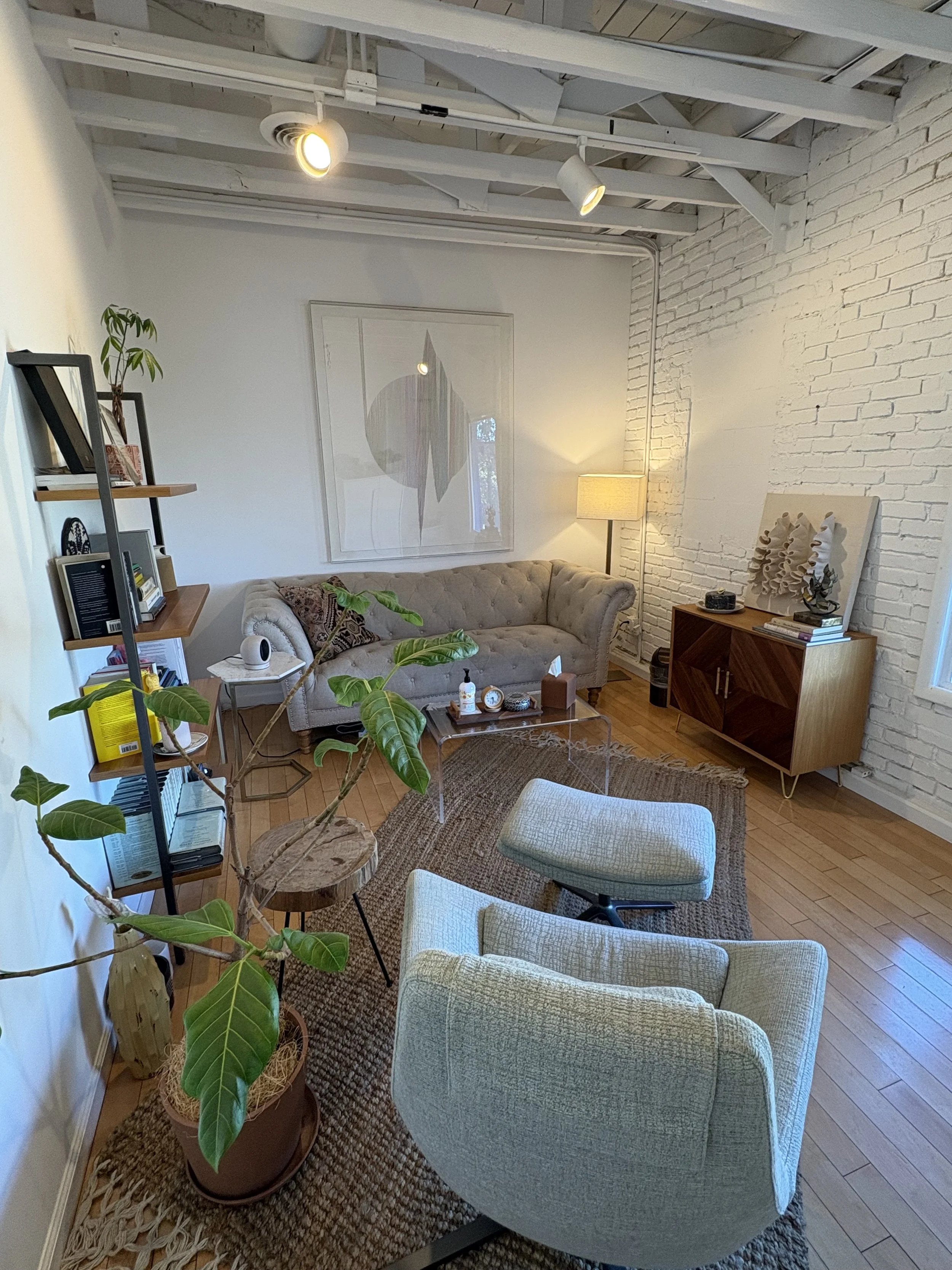 A cozy living room with a beige tufted sofa, two light-colored armchairs, a glass coffee table, a tall lamp in the corner, a potted plant, bookshelves, and artwork on the white brick wall.