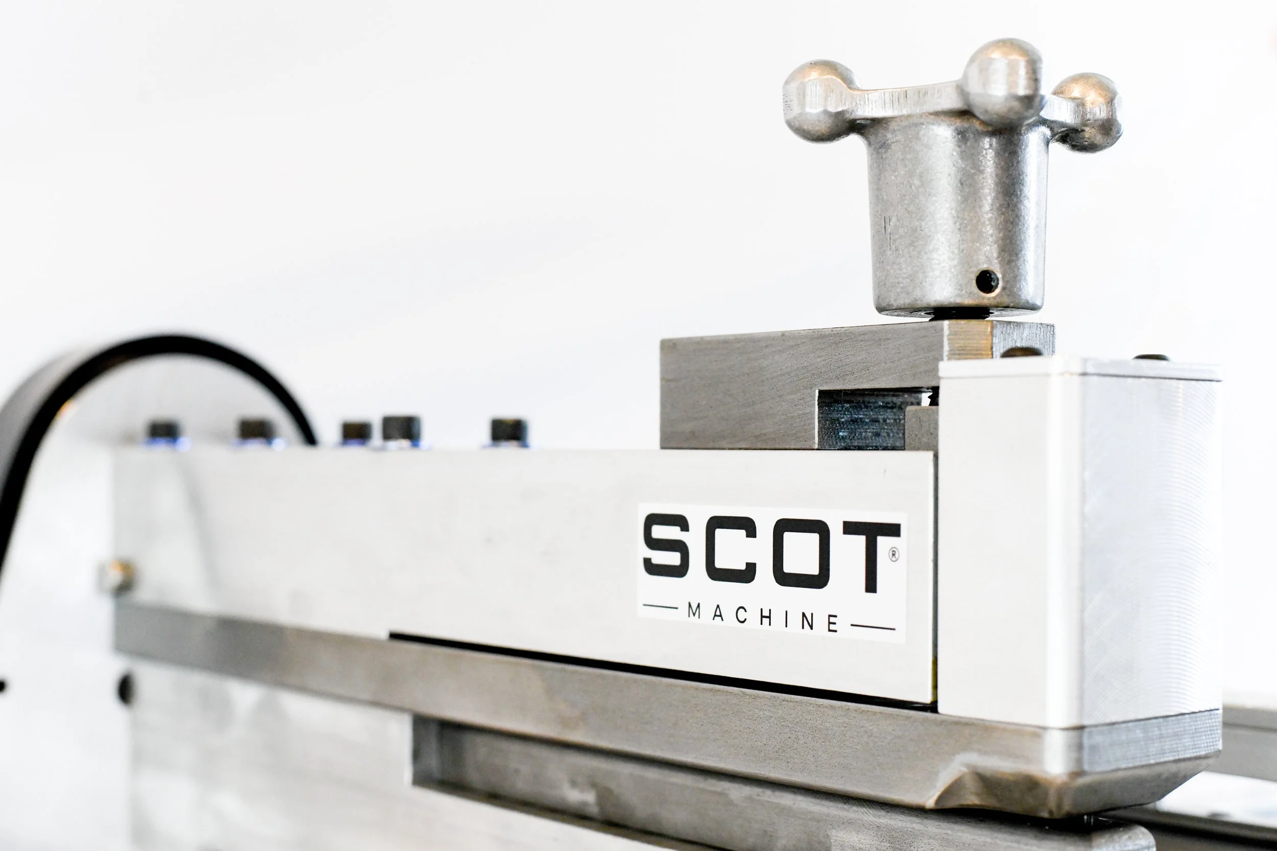 A close-up of a stainless steel machine with the word 'SCOT' visible on a white label, featuring metallic components and knobs, used in industrial or manufacturing settings.