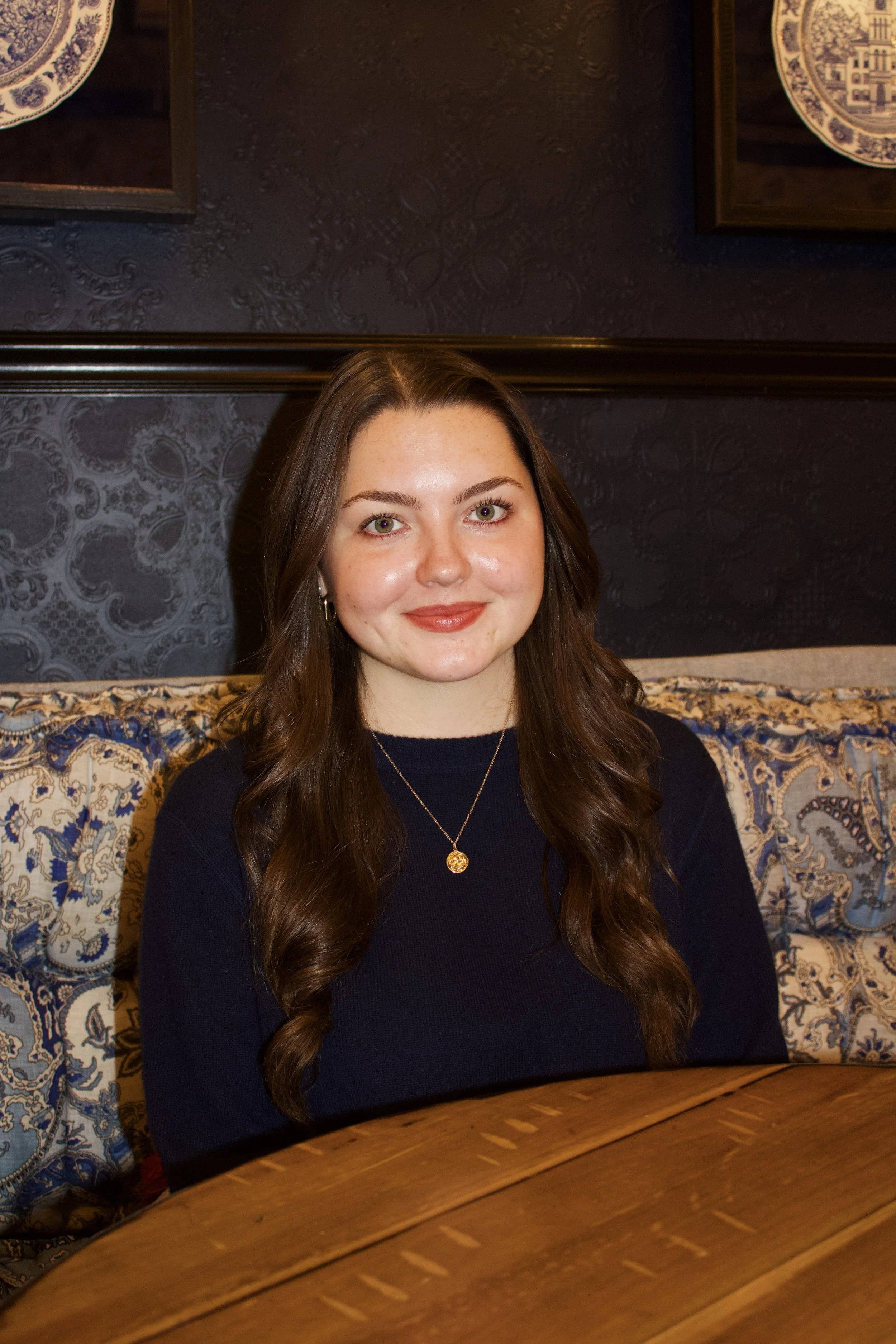 Grey Battle is a young woman with long brown hair and light skin. She sits at a wooden table in front of a dark, textured wall with artwork. She is smiling, wearing a navy top, and a gold necklace with a circular pendant.