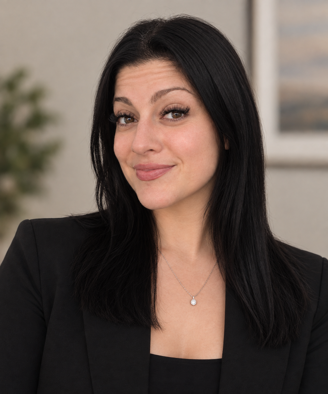 A woman with long black hair, wearing a black blazer and a silver necklace, smiling at the camera indoors.