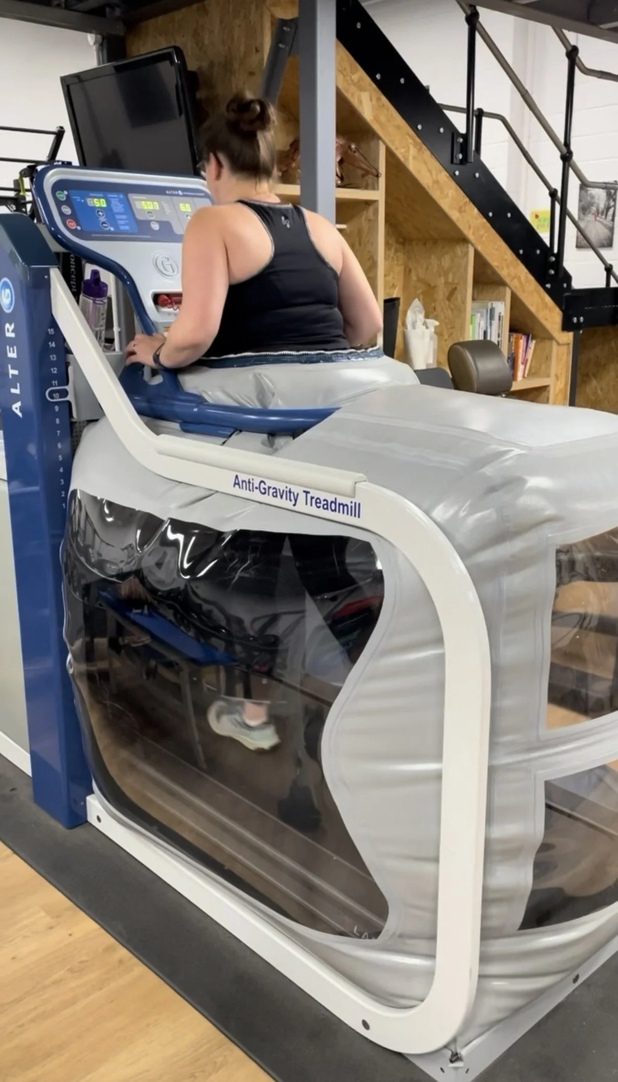A woman using an anti-gravity treadmill, facing away, in a gym or therapy centre. The treadmill has a transparent enclosure and a control panel in front of her.