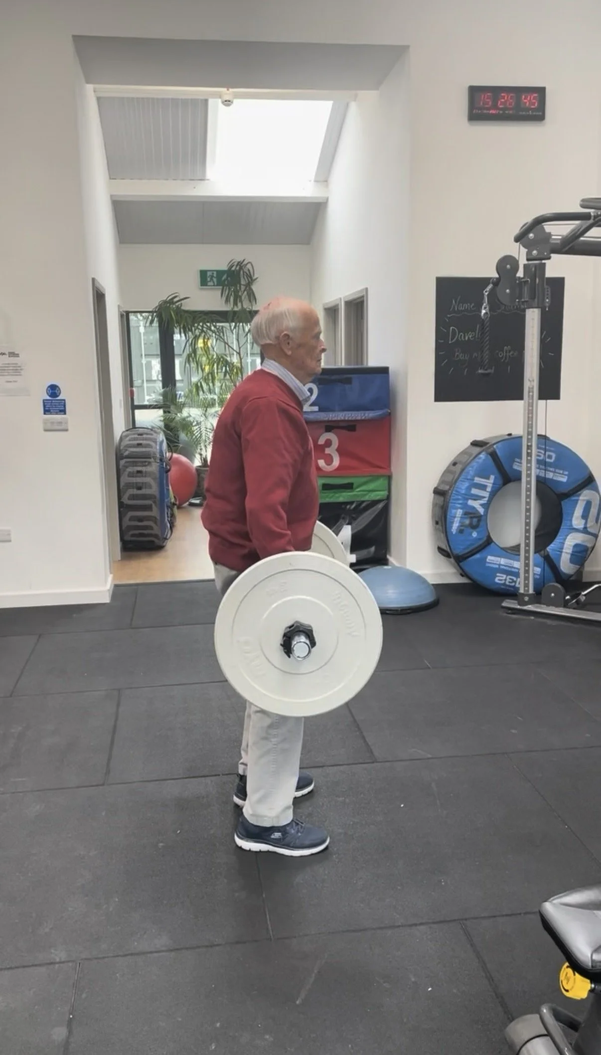 An elderly man standing in a gym holding a barbell with weights, wearing a red jacket, beige pants, and gray athletic shoes. The gym has fitness equipment, a blackboard, and a digital clock on the wall.