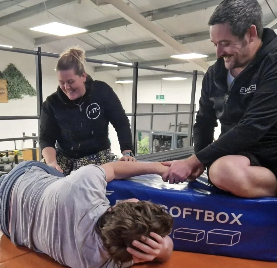 Three people at a gym, with one person lying on the cushioned edge of a padded training box, holding their head with one hand, and two others, a woman and a man, smiling and holding the person's arm during a fitness or training session.
