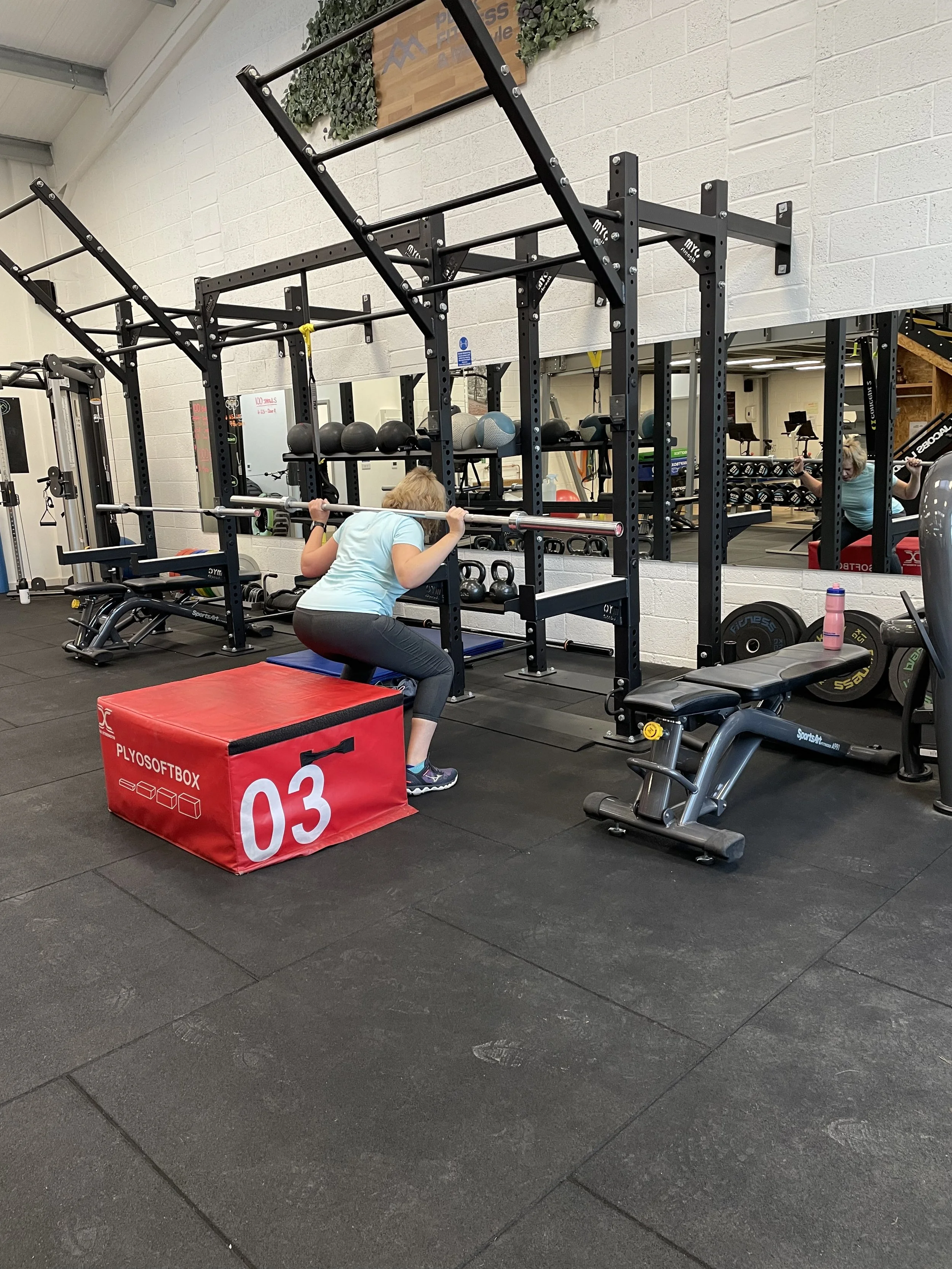 A gym workout area with black metal racks, kettlebells, medicine balls, and a mirror. A woman is squatting with a barbell, and a red plyo box is in the foreground.