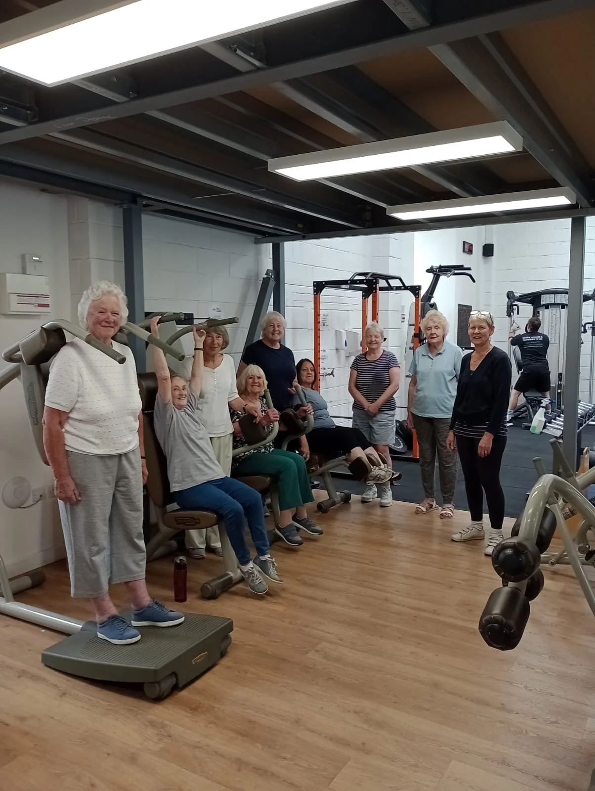 Group of senior women in a gym, some sitting on exercise machines, others standing, smiling, in a workout class.