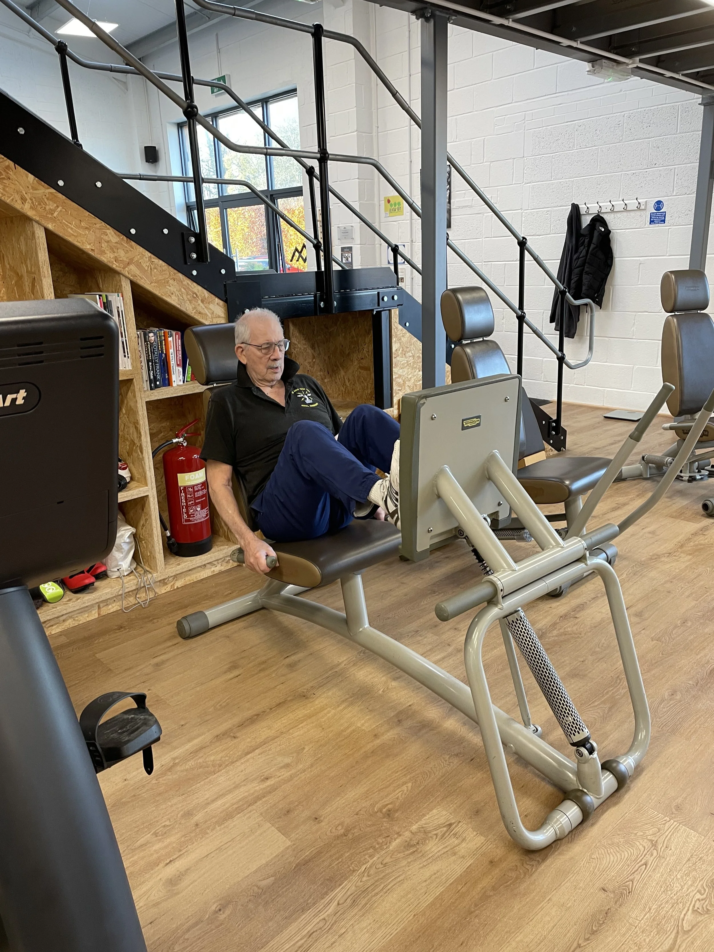 An elderly man with white hair and glasses performs seated leg exercises on a gym machine in a fitness center. The gym has wooden flooring, workout equipment, and a staircase with black railing in the background.