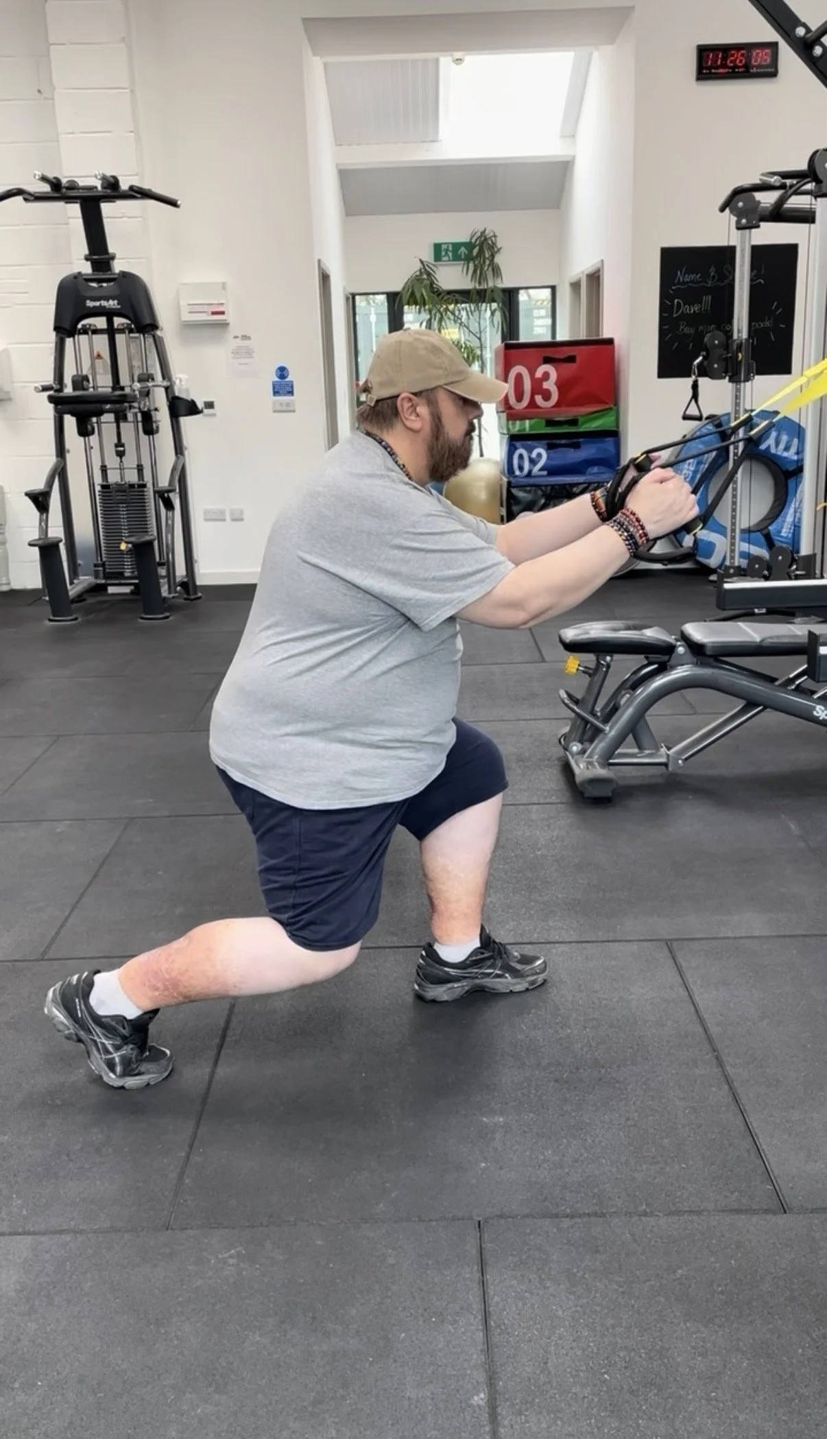 A man in a gray t-shirt, black shorts, and a beige baseball cap is performing a lunge exercise with resistance bands in a gym. The gym has fitness equipment, a blackboard, and a digital clock displaying 11:26.